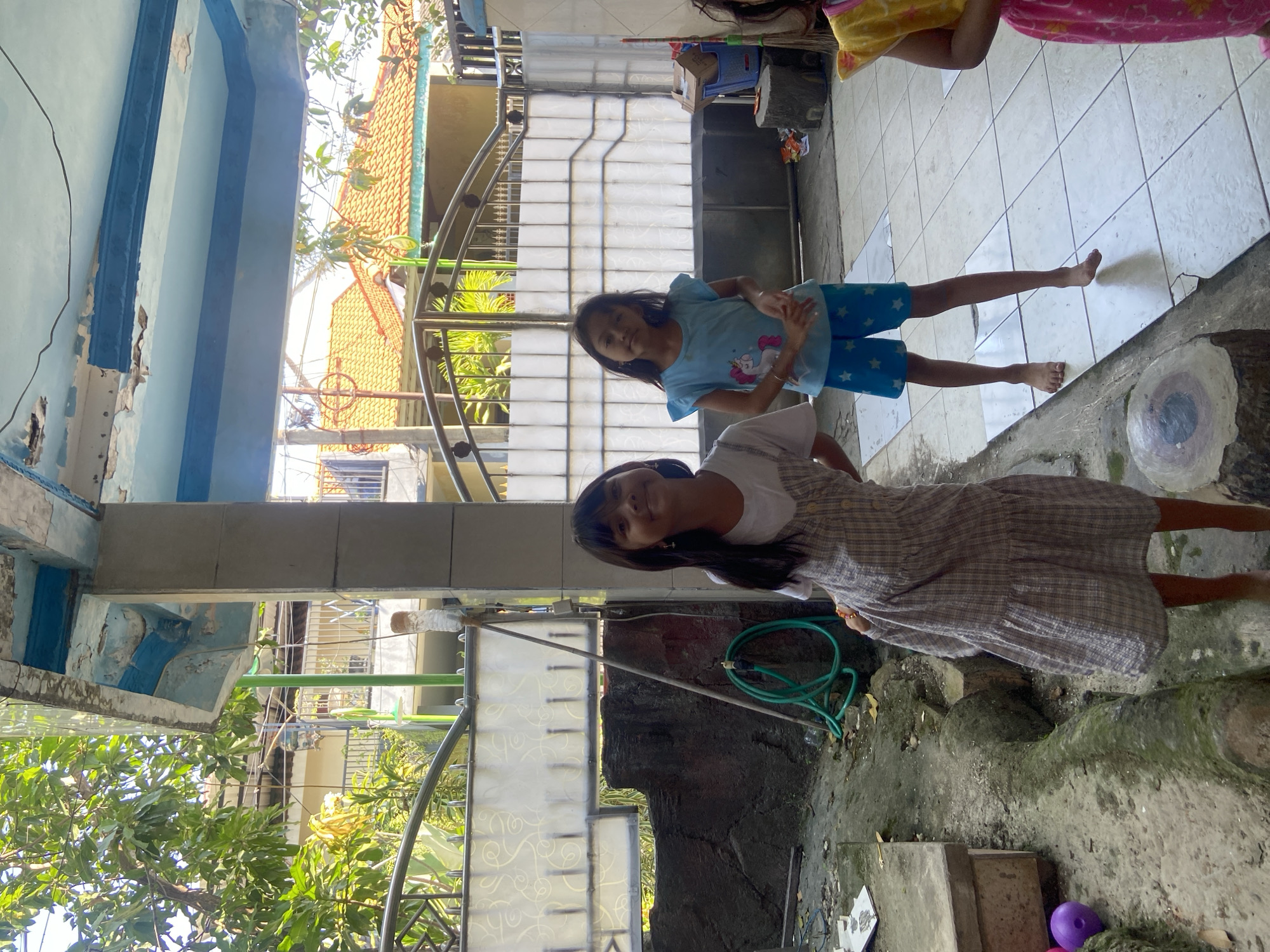The image depicts an outdoor scene in the city of Gelam, Indonesia. In the foreground, two young girls are standing. The girl on the left is wearing a plaid dress over a white t-shirt and is looking at the camera with a smile. The girl on the right is wearing a blue t-shirt with a unicorn graphic and blue shorts with white stars, and she is also looking towards the camera. Behind them, a metal gate with patterned translucent panels is visible, beyond which are residential buildings with orange tiled roofs. The ground is a tiled patio area in front of a weathered concrete structure. Lush green trees and foliage are present in the background. To the left of the girl in the plaid dress, a green hose is coiled on the ground near a dark, textured wall. A purple ball is also visible on the ground in the lower left quadrant of the image. The overall environment suggests a residential area with a somewhat unkempt backyard.