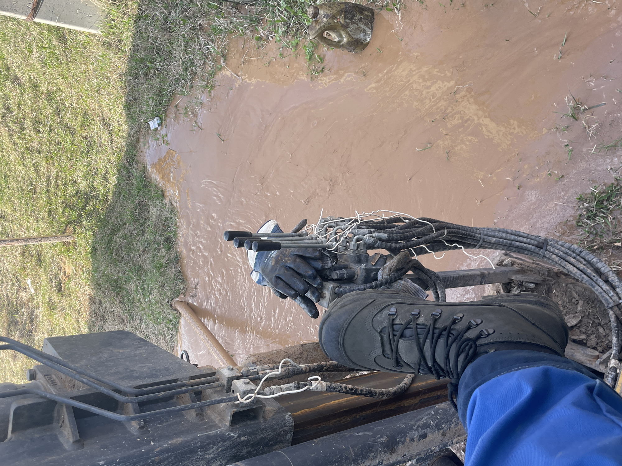 Observed in an outdoor environment is an individual wearing a blue work suit and dark work boots, standing on a piece of heavy machinery. The person is operating the controls of the machine, as evidenced by their gloved hands. Adjacent to the machinery, there is a pool of muddy water. The surrounding environment includes grass and vegetation. Given the muddy conditions and the heavy machinery, the location appears to be a construction or work site. The image does not contain enough information to confirm the location as Поедуги, Russia.