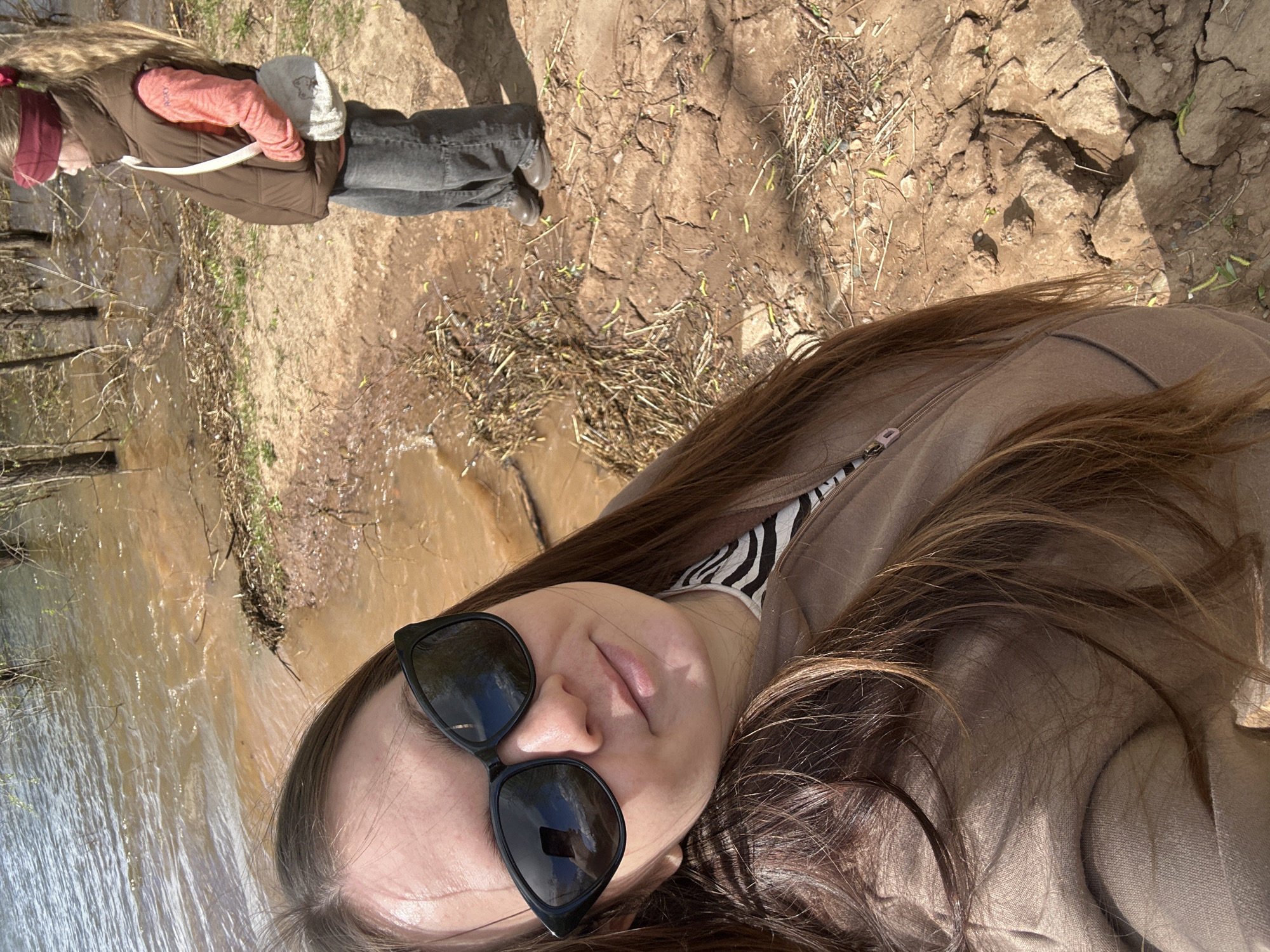 A woman wearing sunglasses and a brown jacket is taking a selfie near a body of water. Another person, wearing a brown jacket, grey pants, and a red hat, stands nearby. The location appears to be an outdoor area with cracked earth and some vegetation. The water is a muddy brown color. The location may be in the city of Chaikovsky, Russia.