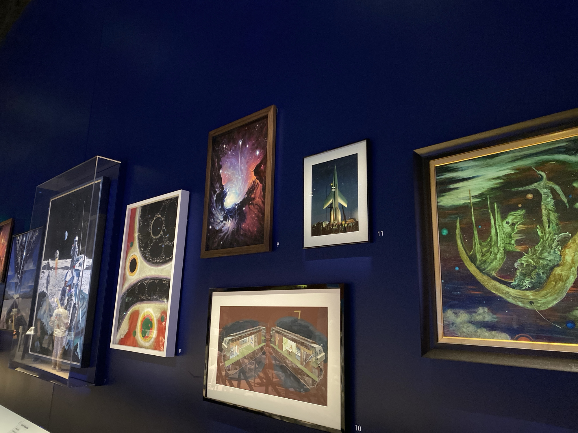 This is a medium-shot, eye-level, indoor photograph of an art exhibition. The exhibition features several framed paintings and prints displayed on a dark blue wall.
On the far left, partially visible, is a large framed artwork depicting a lunar landscape with a spacecraft and an astronaut. To its right is a tall, narrow, framed artwork with a white border, featuring a circular, abstract design with vibrant colors. Next to this is a framed painting with a dark wooden frame, showing a nebula with a starburst in a vibrant display of pinks, purples, and oranges. To the right of that is a smaller, framed print with a white border, depicting a tall, green rocket ship pointing upwards against a dark sky. To the right of the rocket print is a larger framed painting with a dark, ornate frame, featuring a surreal landscape with green, organic forms, possibly alien structures, in a cosmic setting with blue and orange celestial bodies.
Below the rocket ship print and to the left of the surreal landscape painting is a horizontally oriented framed artwork with a dark frame, showing two cross-sections of what appear to be living spaces, possibly train cars or futuristic dwellings, with interior details visible. Numbers are subtly placed near some of the artworks, suggesting a cataloging system (e.g., "8", "9", "10", "11").
The lighting is dim and focused on the artworks, creating a dramatic effect. The overall impression is that of a museum or gallery space dedicated to science fiction or space-themed art. The location is likely within the city of Washington, United States of America, given the context of an art exhibition.