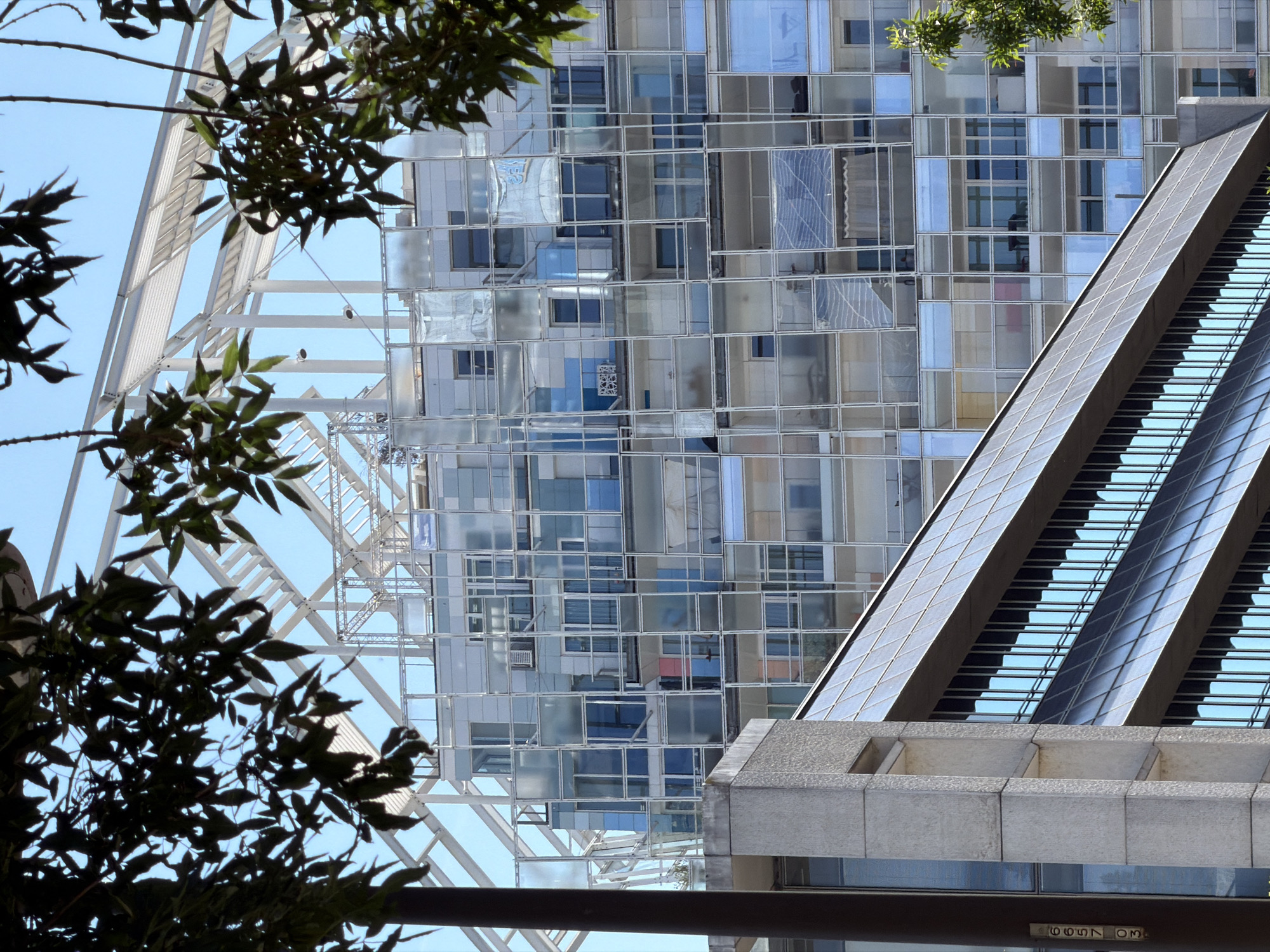 The image captures a segment of the Confluence district in Lyon, France, an area known for its modern architecture. In the foreground, a concrete and glass building with a sloping, dark-tiled roof is visible. The building has multiple floors with rows of reflective glass windows that mirror the blue sky. To the left, a vertical column with numbers "6657 03" is mounted on a textured concrete facade.
Behind this structure, a more complex, multi-story building with a distinctive façade composed of numerous glass panels and balconies is prominent. The glass panels vary in transparency and tint, creating a mosaic effect. Some panels reveal glimpses of the interior, showing balconies, furniture, and what appear to be curtains or screens. The building's structure is a grid of slender white metal frames.
Above and behind these buildings, a white, skeletal architectural structure with visible beams and supports is seen against a clear blue sky. This structure appears to be a large, open framework, possibly part of a larger building or an installation. Dark green foliage from trees partially obscures the view of the buildings and sky, framing the top and right side of the image. The overall impression is one of contemporary urban design with a focus on glass and metal.
