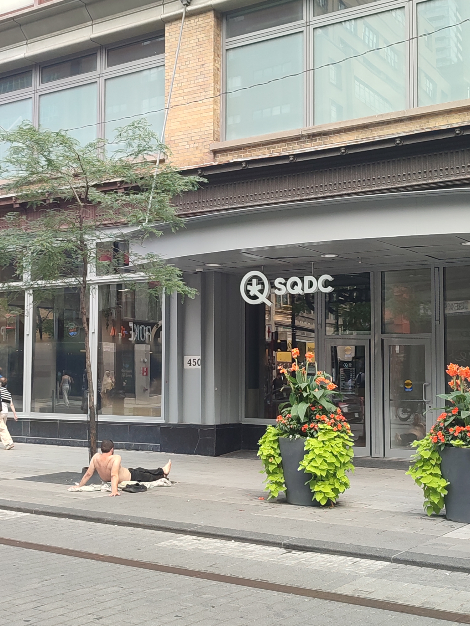 An outdoor shot taken on a bright day in Montreal, Canada, features a man shirtless and lying on the sidewalk in front of a building with storefronts. He is positioned on a towel or blanket with his legs extended and appears to be resting. A tree with green foliage stands to his left. The building has large windows and a prominent sign that reads "SQDC." Two large planters filled with vibrant green and orange flowers are placed near the entrance of the building. The sidewalk is paved with gray stones, and a street with tram tracks runs in the foreground. The overall environment suggests an urban setting with commercial establishments.