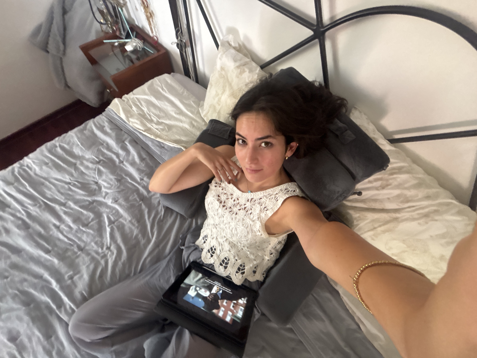 A young woman is lying in bed with a black tablet in her lap. She is wearing a white, crocheted sleeveless top and gray pants. She is propped up by gray pillows. Her right arm is extended towards the camera, revealing a gold bracelet. The bed has gray and white bedding. In the background, there is a black metal bed frame, a white wall, a wooden bedside table, and some decorations. There is no indication of the location being in the city of Puebla, Mexico, or any other specific location.
