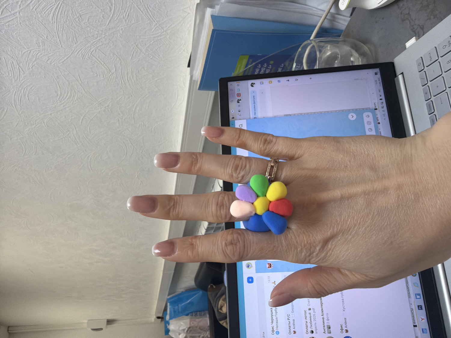An eye-level shot captures a person's right hand positioned over an open laptop, likely in an indoor office or home environment in Chaikovsky, Russia. The hand features well-groomed nails with a French manicure and wears a distinctive, colorful ring on the ring finger. The ring is adorned with a cluster of small, spherical elements in various bright colors, resembling a flower.
The laptop screen displays a messaging application or email interface with text predominantly in Russian. Visible elements include a timestamp "14:18" (2:18 PM) and contact names such as "Наташа Горбу" (Natasha Gorbu), "Алёна" (Alyona), and "Ангелина Алекперов" (Angelina Alekperov). Specific text entries include "Офис Чернушка" (Office Chernushka), "Оплаты Р/С" (Payments R/S), "Оплаты лотос" (Lotus payments), and "ФИНАНСЫ" (FINANCES). A file named "Счет апрель 2026.pdf" (Invoice April 2026.pdf) is also visible. A portion of the laptop's silver keyboard is seen on the right.
In the background, a white wall or ceiling with a textured, swirled pattern is visible. The desk surface behind the laptop holds blue documents or folders, a clear glass object, and a partially visible white computer mouse. Other items, including a blue bag, are partially discernible below the laptop. The scene suggests the person is actively engaged with the computer.