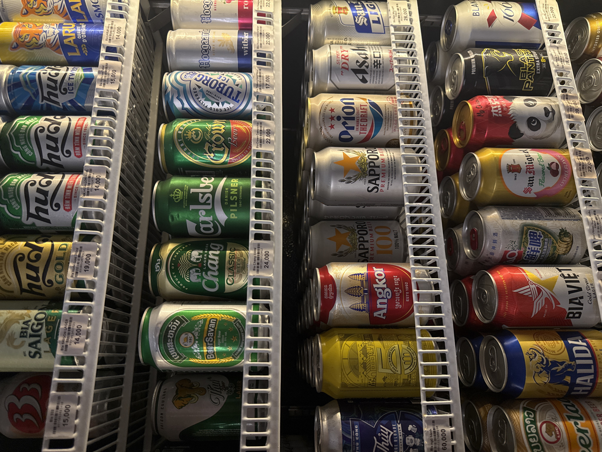 The image displays a refrigerator filled with a variety of canned beverages, primarily beer, arranged on shelves. The refrigerator is likely located in a convenience store or supermarket in Đà Nẵng, Vietnam, given the presence of Vietnamese product labels and pricing in Vietnamese Dong. The products are neatly organized, with price tags visible on some of the shelves. The lighting suggests an indoor setting, possibly a brightly lit retail environment. There are no people visible in the image.