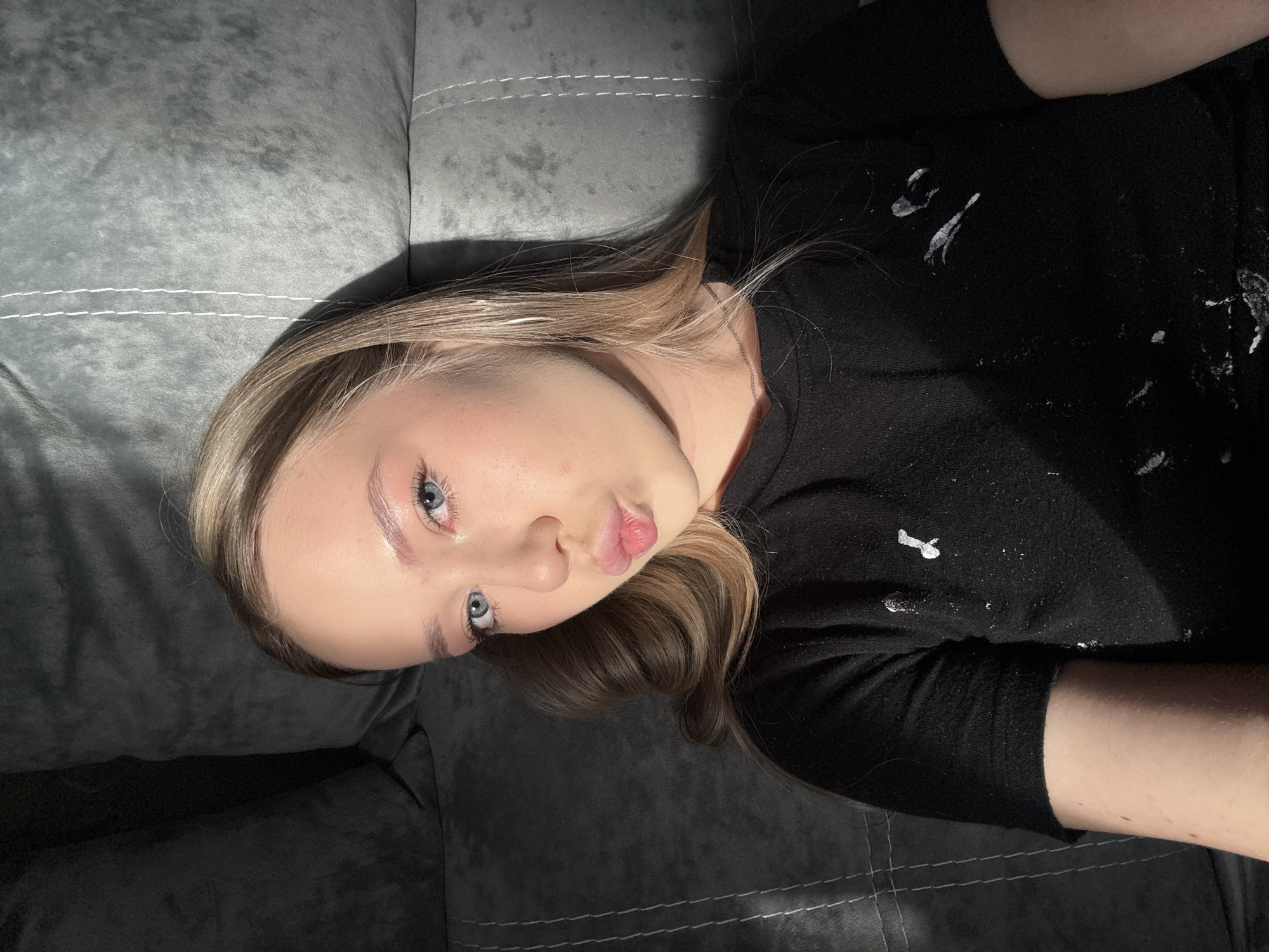 A young woman with light blonde hair and blue eyes lies on a gray upholstered surface, likely a couch or bed. She is wearing a long-sleeved black shirt that appears to have some white paint or similar substance on it. Her lips are slightly pursed. The lighting is uneven, creating strong shadows on the left side of her face and the upholstered surface. The photograph appears to be taken from a slightly high angle, as if the photographer is looking down at her. There is no information to confirm the location in the city of Chaikovsky, Russia or any other specific location.