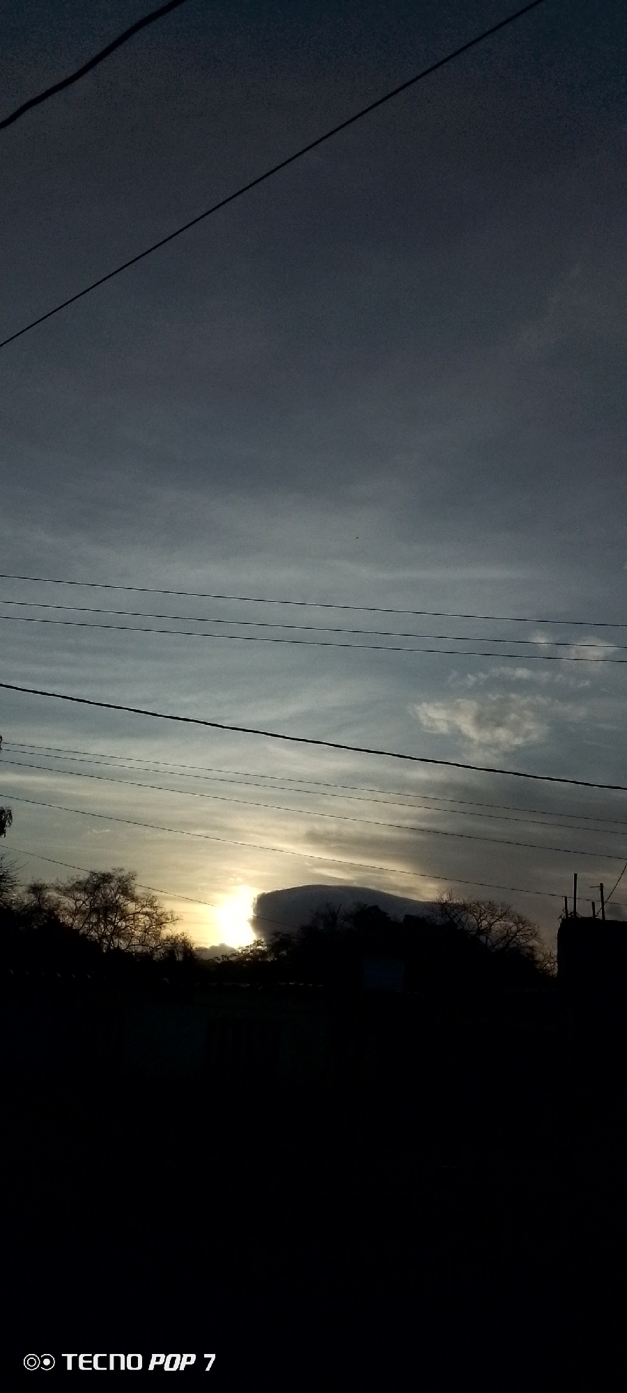 The image shows a landscape with a partially obscured sunset in Altagracia, Venezuela. Silhouetted trees and buildings are in the foreground, with electrical wires stretching across the upper part of the frame. The sky is a gradient of light and dark grey, with thin clouds near the horizon where the sun is setting. The lower part of the image is dark, making details in that area difficult to discern. A "TECNO POP 7" watermark is visible in the lower-left corner.