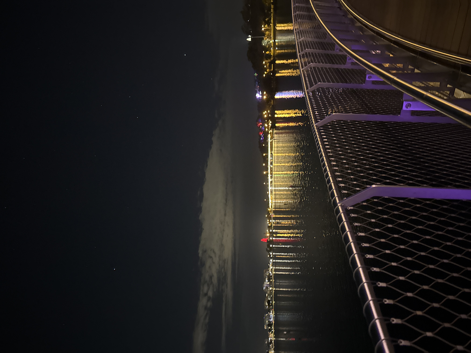 This night scene captures a modern bridge or elevated walkway curving gracefully over calm water in Perth, Australia. The perspective is taken from the walkway itself, which occupies the lower right portion of the frame, featuring sleek metal railings and a textured, possibly wire mesh or grated surface. Sections of the walkway are subtly illuminated with a soft purple glow, creating an atmospheric effect.
To the left of the walkway, the water reflects a vibrant array of artificial lights from the distant shoreline, appearing as shimmering vertical streaks of yellow, white, red, and blue. Across the water, the distant shore reveals a lively cityscape or town, densely dotted with lights from buildings and structures.
The night sky dominates the upper left, a dark expanse dotted with a few faint stars. A long, thin band of clouds stretches horizontally across the sky above the water, adding a touch of natural texture to the dark heavens. The overall mood is one of urban tranquility and illuminated beauty under a starry, cloudy night. No people or visible text are present in the image.