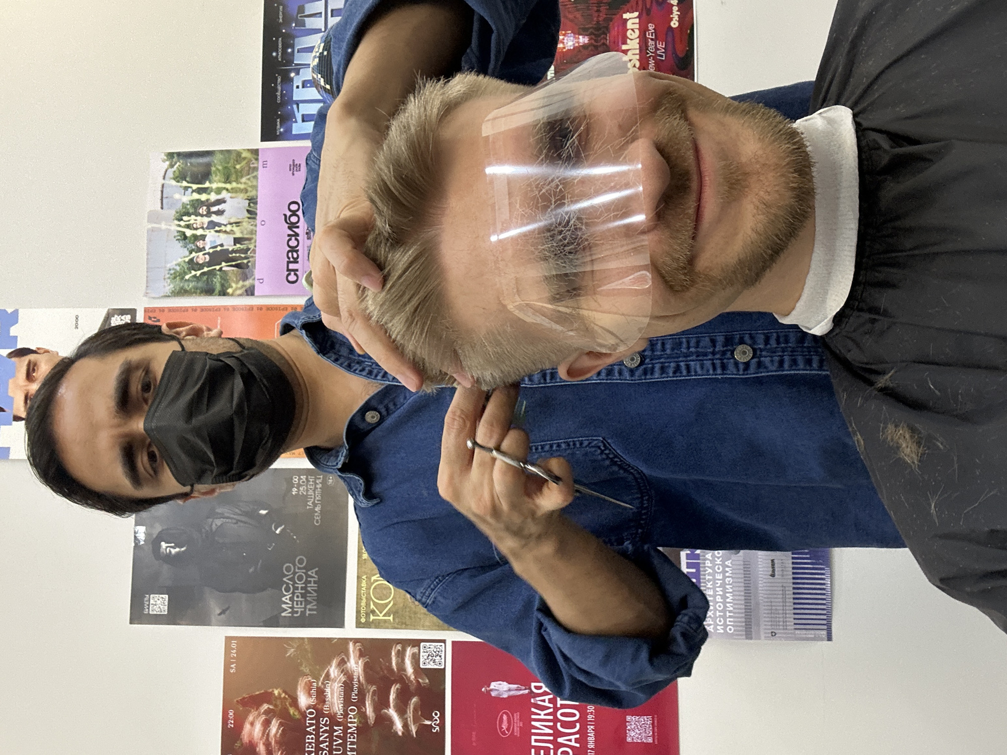 A man with a black face mask is giving a haircut to a man wearing a clear plastic face shield. The location appears to be a hair salon in Tashkent, Uzbekistan, evidenced by the posters in the background. The barber is holding a pair of scissors and is trimming the hair of the customer. The customer is wearing a black cape with a white collar. The posters on the wall are advertising various events and exhibitions, including a photography exhibition, an architectural event, and concerts.
