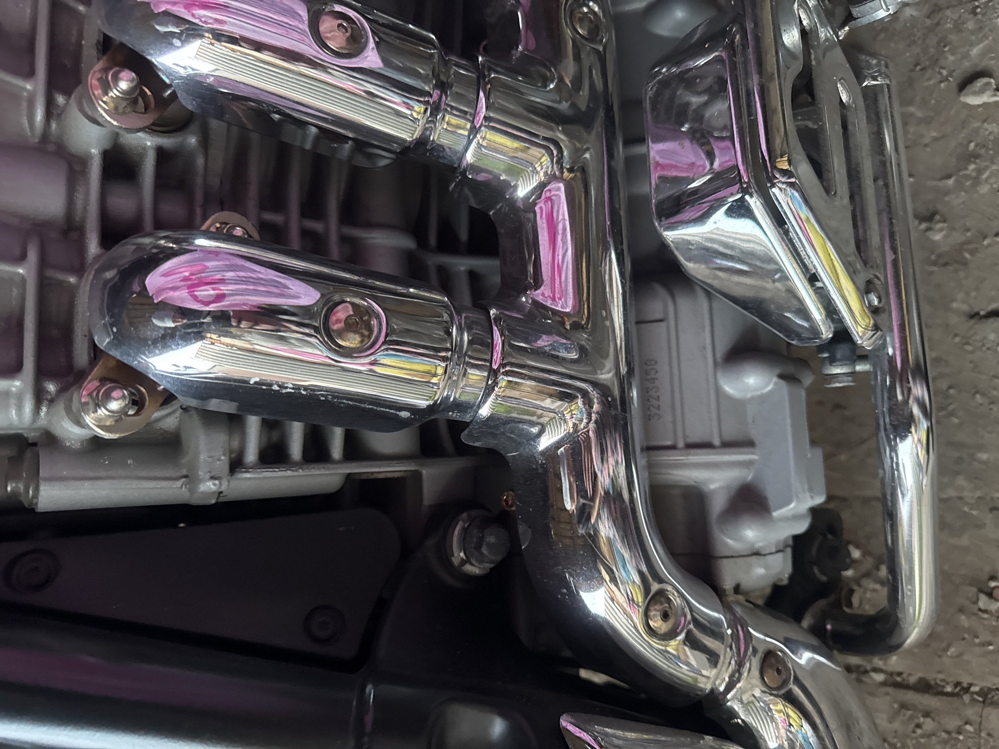 A close-up, eye-level, daylight, medium shot captures the chrome exhaust pipes of a motorcycle engine. The chrome reflects a pinkish hue and the surroundings, including what appears to be a blurry person in the reflection of the upper pipes. A grey engine block is visible behind the pipes, with a serial number that reads "3223458". The motorcycle is situated on a ground surface that appears to be concrete or asphalt, with a slight texture. The scene is set in Боголюбово, Russia.