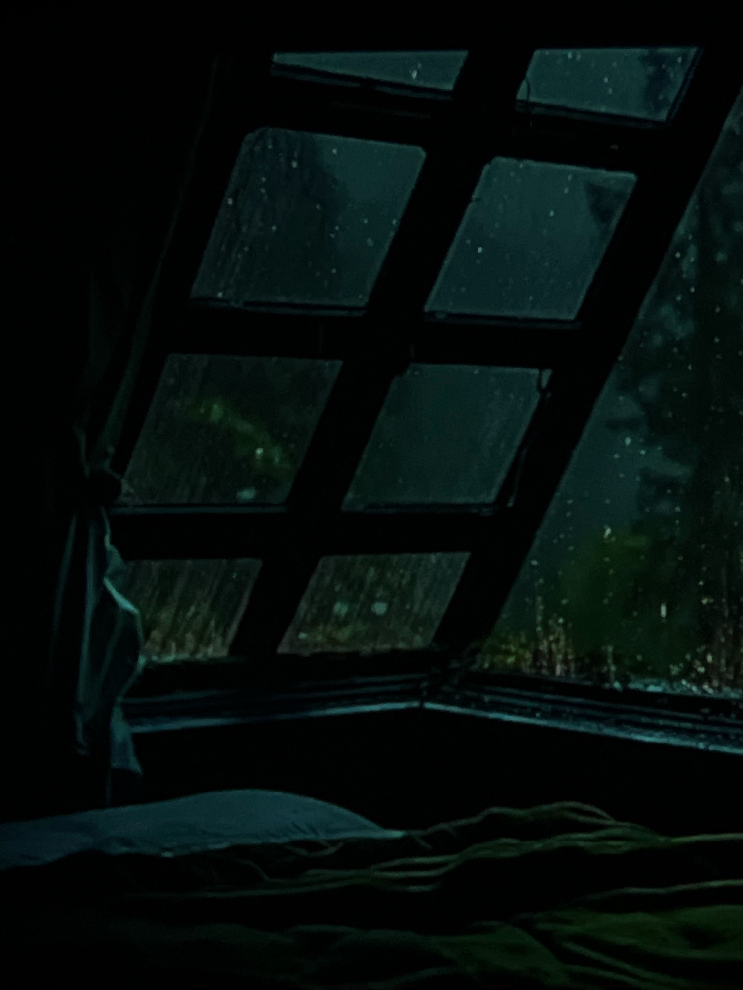 The image depicts an interior view looking out of a window, suggesting it might be a bedroom or a sleeping area. The window is divided into multiple diamond-shaped panes, and it appears to be raining outside, with streaks of water visible on the glass. The exterior scene is dark and obscured by the rain and low light conditions, but hints of trees and foliage can be discerned. Inside the room, a bed with blankets and pillows is visible in the foreground, covered in dark green and blue fabric. A white cloth or curtain hangs loosely on the left side of the window frame. The overall atmosphere is dim and somewhat melancholic due to the rainy weather and low lighting. The location in the city of Wałcz, Poland is not identifiable from the visual information provided in the image.