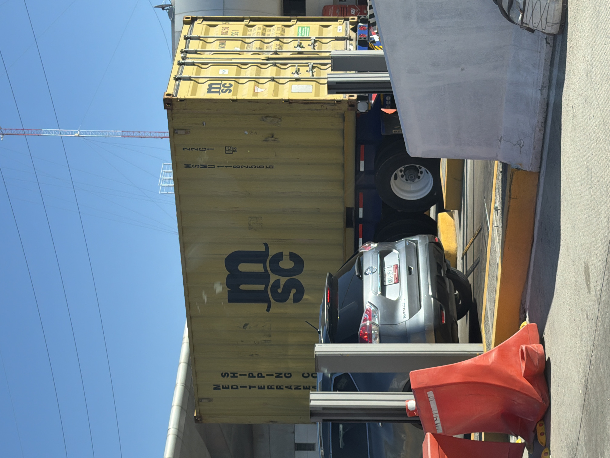 The image displays an outdoor scene in Santa María Moyotzingo, Mexico, under a clear blue sky with multiple power lines visible overhead. A large, yellow corrugated MSC shipping container is centrally positioned, marked with "MSC" logo, "MSMU 182565," "MSMU 18256," and "MEDITERRANEAN SHIPPING CO." This container is mounted on a dark blue truck trailer, exhibiting white-rimmed wheels. In front of the container, a silver Nissan X-Trail SUV, with a Mexican license plate reading "VMS-351-A," is partially obscured by two gray rectangular metal barrier posts. A second, darker-colored vehicle is partially visible to the left of the SUV. The immediate foreground contains two red plastic traffic barriers on paved ground, bordered by yellow painted curbs. A substantial light gray concrete barrier is positioned on the right, partially obscuring the truck trailer. In the background, a tall, red and white striped antenna tower is visible, along with a curved light-colored overhead structure on the upper left. No human individuals are present in the image.