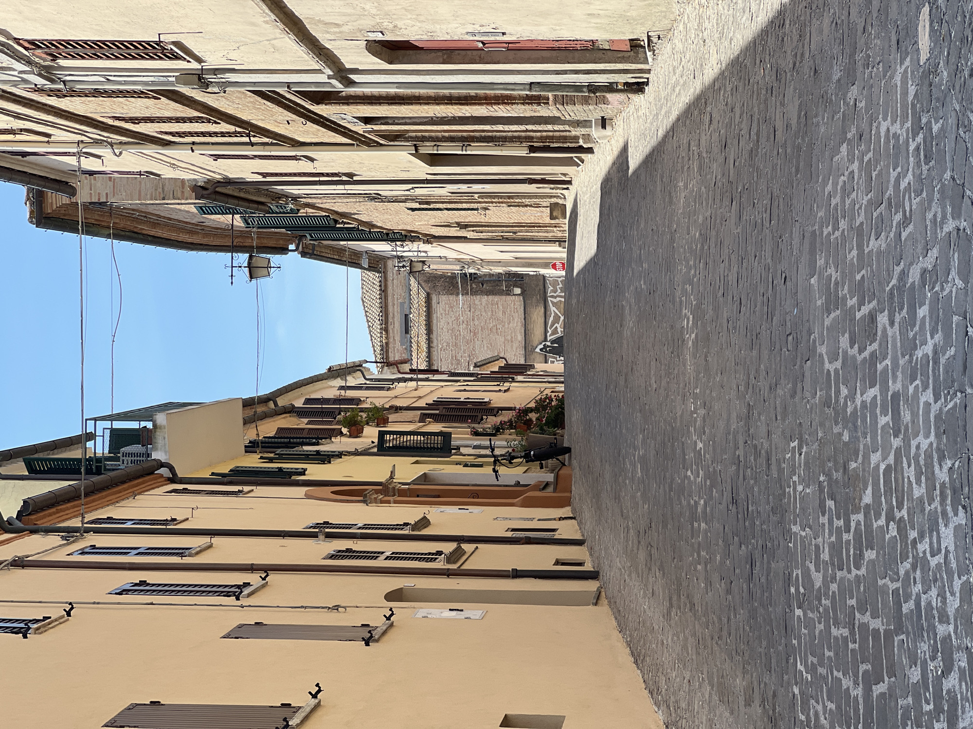 The image depicts a narrow, upward-sloping cobblestone street in Barbara, Italy, lined with buildings on both sides. The buildings are constructed of brick and plaster, with facades painted in muted tones of yellow, beige, and ochre. Numerous windows with dark wooden shutters are visible. On the left side of the street, a building with a peach-colored facade features several windows and a small recessed doorway. On the right side, buildings display exposed brickwork and darker painted doorways. Hanging wires are strung across the street overhead. A bicycle is parked near the right-hand wall midway up the incline, next to potted plants. Further up the street, a red stop sign is visible on the right side. The sky is clear and blue. The sunlight casts strong shadows across the cobblestones, indicating a sunny day. The overall impression is one of a quiet, historic European street scene.