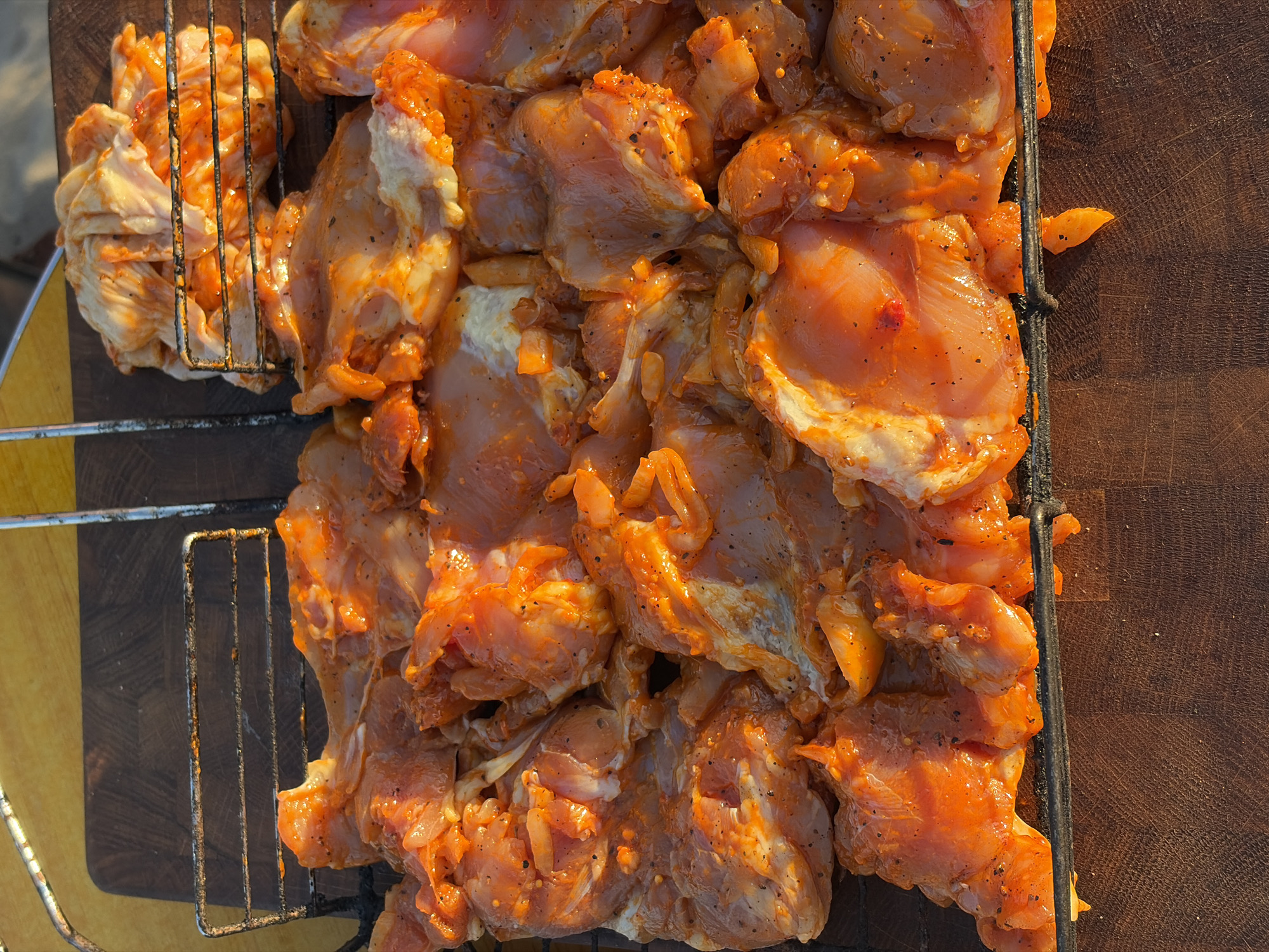 The image shows a close-up, overhead view of raw chicken pieces marinated in an orange sauce, arranged on a metal grill rack. The chicken is seasoned with black pepper. The grill rack is positioned on a dark wooden surface, possibly a cutting board or table. In the background, another grill rack with more marinated chicken is visible, stacked higher. The lighting suggests it is either early morning or late afternoon, casting a warm glow on the scene. The location is described as being in the city of Поваровка, Russia. There are no people or animals present in the image.