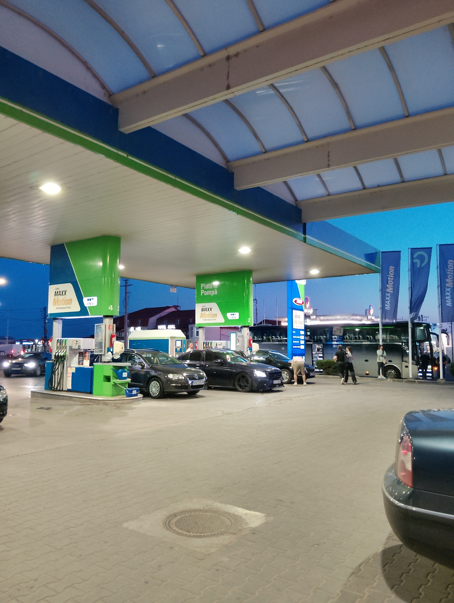 The image shows a gas station in Adjud, Romania. In the foreground, there is a dark-colored car. In the midground, several cars are parked at the gas pumps, and a bus is visible behind them. There are also a few people present, appearing to be customers or employees. The gas station is covered by a canopy with blue and green elements, and illuminated by artificial lights. The sky is dark, indicating it is either dusk or night. The ground is paved with interlocking concrete blocks. Several flags with the "MAXX Motion" logo are displayed.