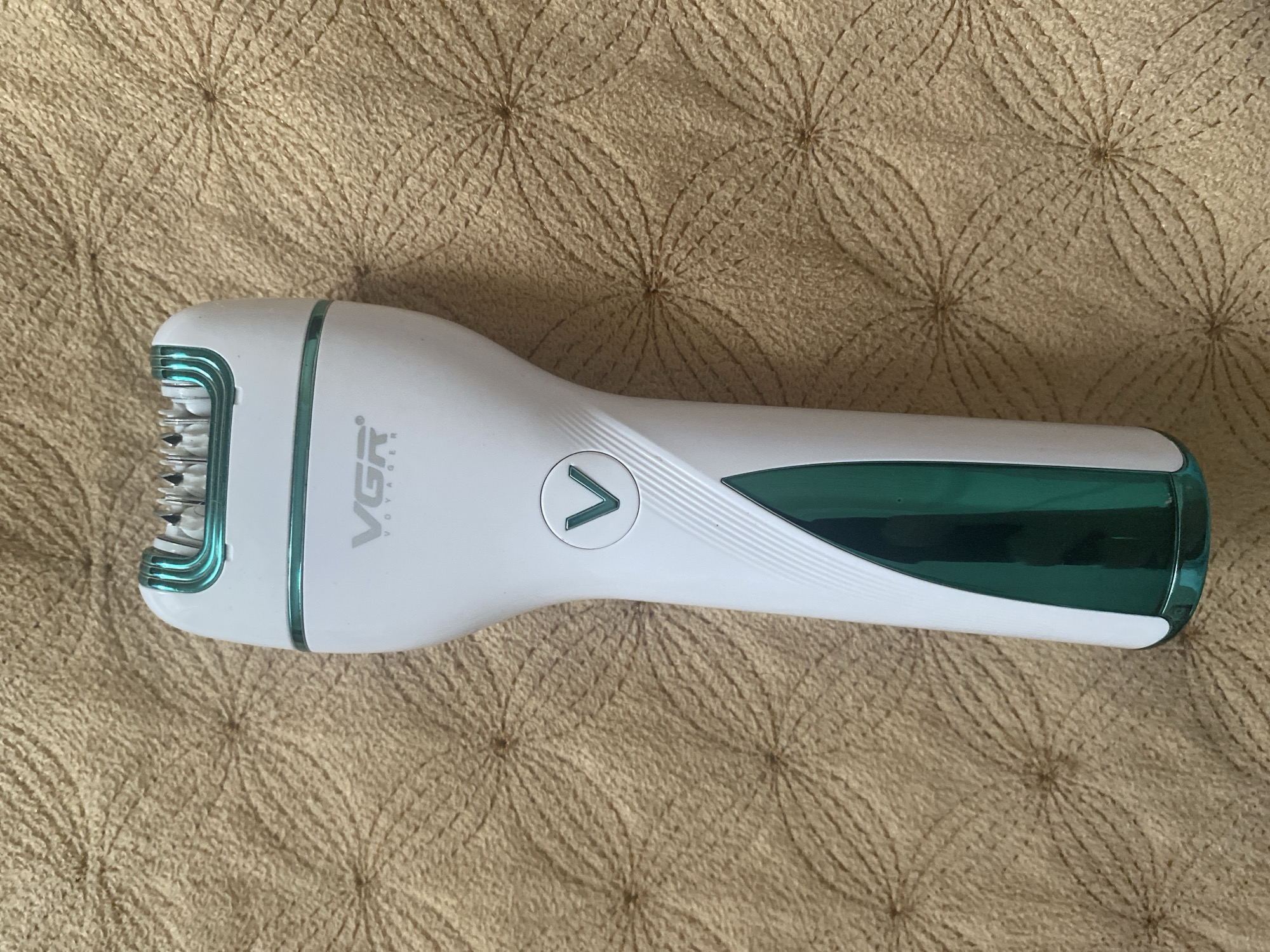 An epilator with a white and green body is laid on a textured brown blanket. The epilator is positioned vertically in the center of the frame, with its top part, which contains the epilating head, facing upwards. The epilating head has several small blades visible. The epilator features the "VGR" logo in black text. Below the logo, there is a green "V" symbol within a circular indentation. A diagonal green insert is present on the lower part of the epilator's body, adding a decorative element. The blanket has a repeating pattern of light brown, circular motifs. The image does not contain any people or indicate a specific city or location within Russia.