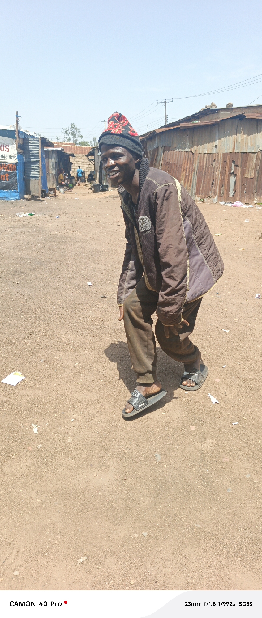 In Barikin Gidar Kanat, Nigeria, a man stands in a dusty, open area, likely a street or market. He is wearing a dark beanie with red lettering, a brown quilted jacket, and brown pants. His right leg is bent, and he is wearing black slide sandals on both feet. He is smiling and looking to his left. The background shows several makeshift structures made of corrugated metal and possibly brick, with some clothing and debris scattered on the ground. The sky is clear and blue.