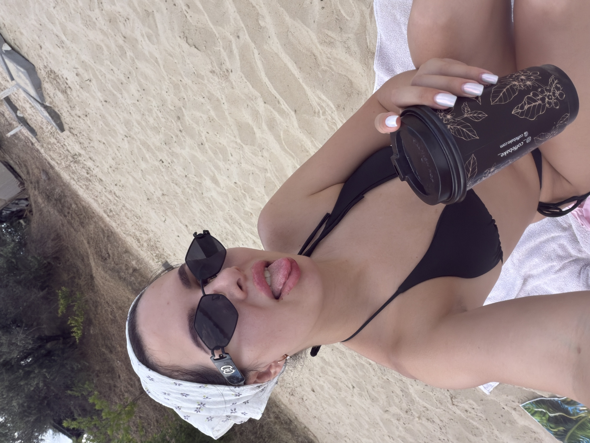 A young Caucasian woman wearing a black bikini, a white bandana, and black sunglasses poses for a selfie on a sandy beach. She is sitting with her tongue sticking out. She holds a black disposable coffee cup with a leaf pattern in her right hand. The background consists of sand, trees, and some beach furniture.