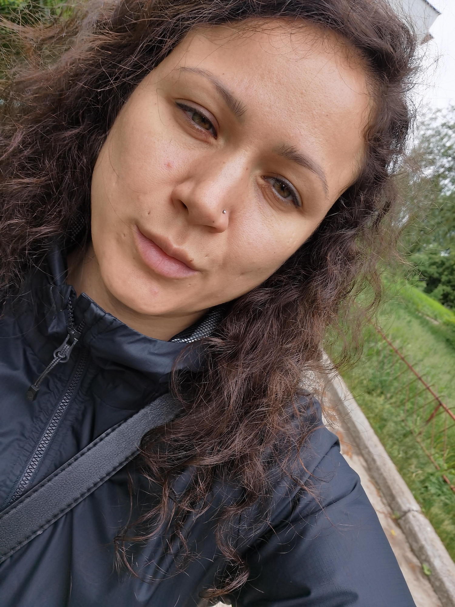 A close-up selfie-style image depicts a female subject with dark, curly hair and medium skin tone, looking directly into the camera. Her facial features include brown eyes with visible eyeliner on the upper lid, naturally dark eyebrows, and a silver-colored stud piercing on her left nostril. Her lips are a natural pink hue and slightly parted. Visible skin texture includes minor blemishes on the forehead and cheeks. She is wearing a dark grey or black zippered jacket, likely made of a synthetic material such as nylon, with a visible zipper down the front and a textured internal collar. A dark, possibly fabric, strap from a bag crosses her upper torso. The background indicates an outdoor setting, with green grass, foliage, and trees visible. A rusty metal fence or railing is partially visible on the right side of the frame, and a concrete curb or edge defines a boundary in the lower right. The sky overhead appears overcast. The image lacks specific identifying markers to confirm the location in the city of Chaikovsky, Russia, beyond generic outdoor elements.