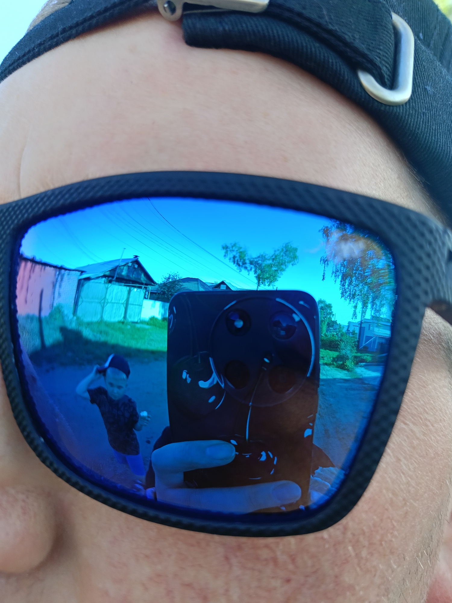 A close-up shot focuses on a person wearing blue reflective sunglasses, revealing a reflection of a neighborhood scene in Chaikovsky, Russia. The reflection shows a boy wearing a black t-shirt and blue shorts walking along a dirt road, partially obscured by the hand holding a smartphone. A house with a light-colored facade and a dark roof is visible in the background, along with a fence and power lines stretching across the sky. The sunglasses wearer is wearing a black cap with a metal clip.