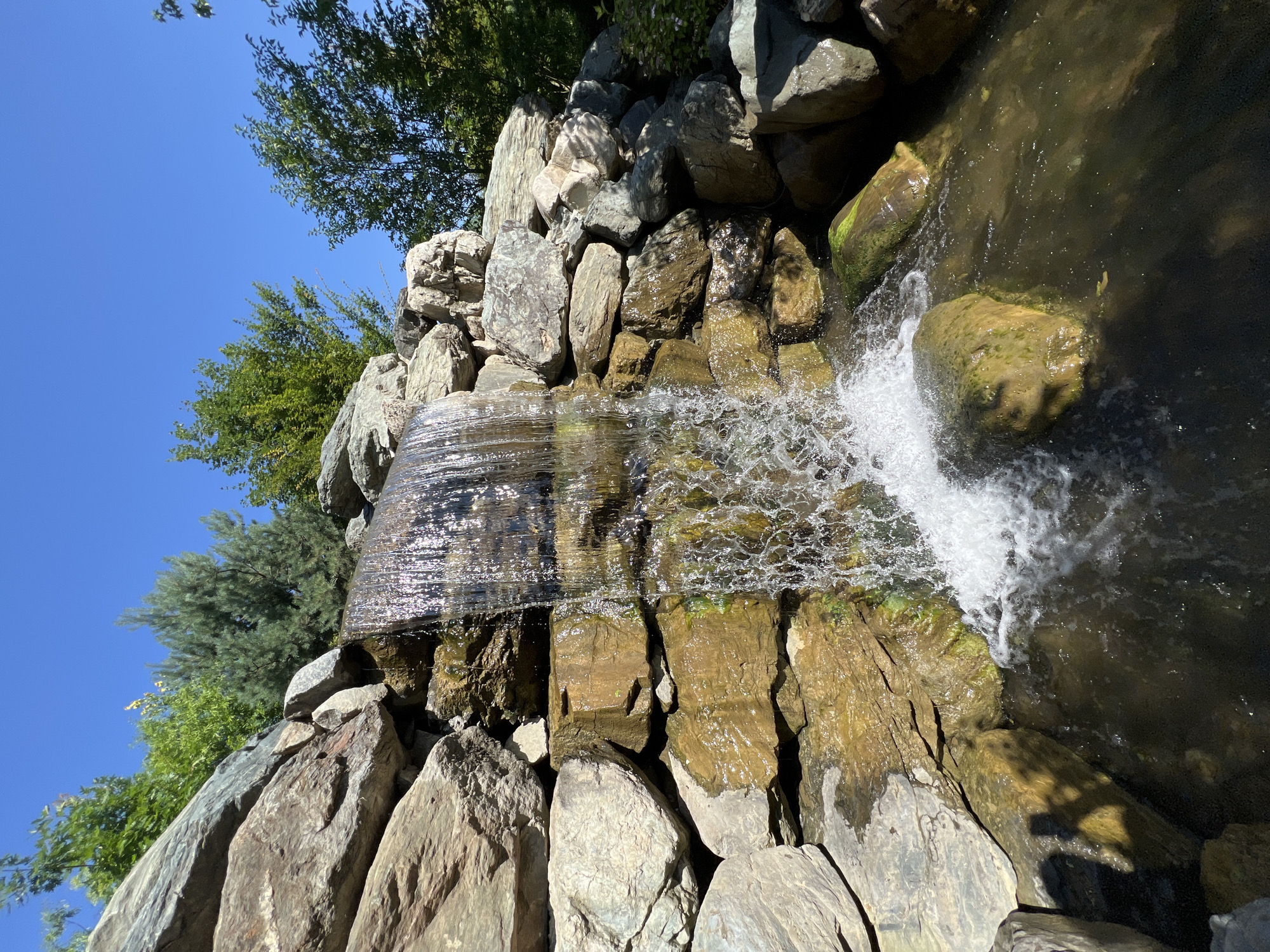 A man-made waterfall cascades over a rocky embankment into a pool of dark water. The rocks are a mix of grey and tan, with some moss and greenery clinging to them. Lush green trees and a clear blue sky are visible in the background. A shadow of a person, presumably the photographer, is cast on a rock in the foreground. The scene appears to be in a park or garden setting.