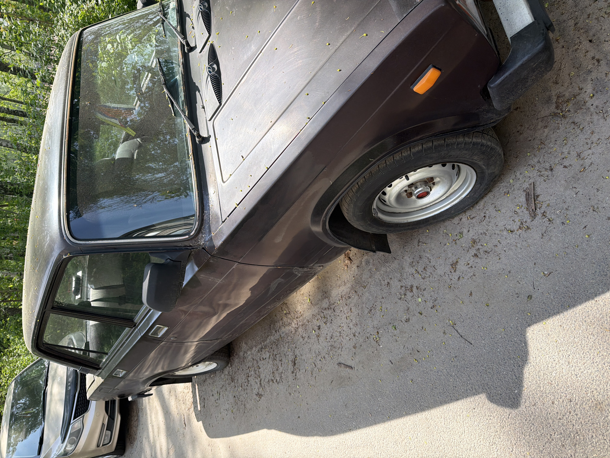 A brown Lada sedan is parked on an asphalt surface. The car is partially obscured by shadow and positioned at an angle relative to the viewer. The passenger-side front tire is visible and appears to be in good condition. The car is covered with yellow pollen.
The location is not discernable as Tokkari-Lend, Russia, based on the image alone. There are trees in the background. Another car is parked to the left of the Lada. The scene appears to be outdoors during daylight.