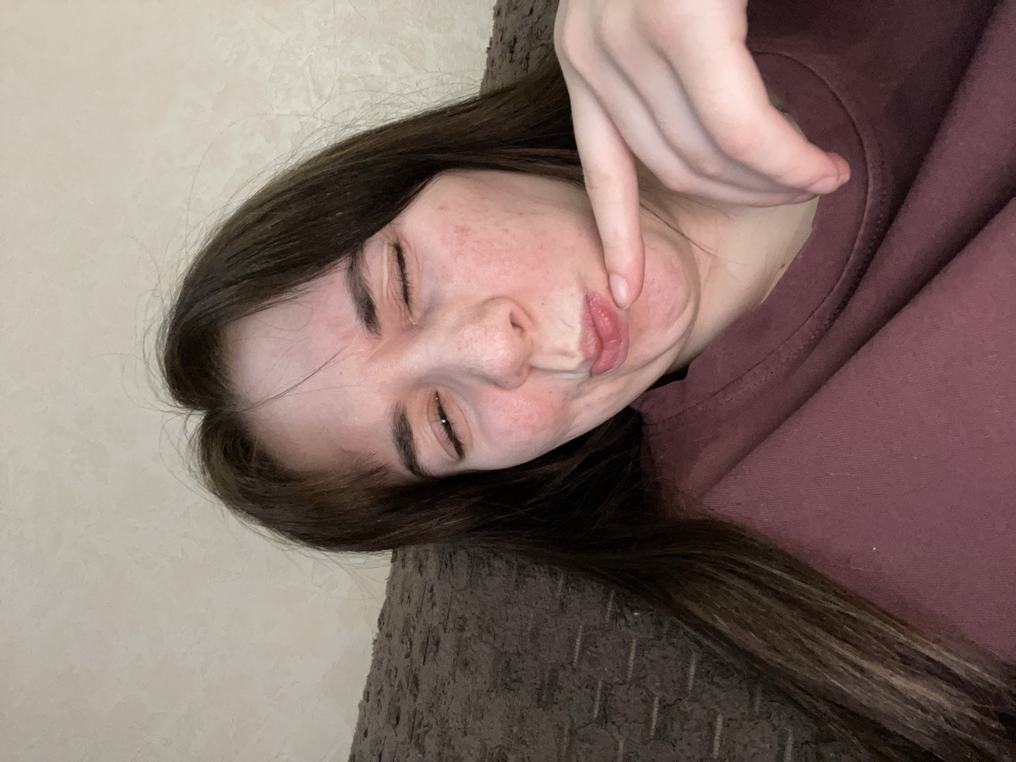 A woman is lying down on a brown textured surface, possibly a couch or bed. She is wearing a maroon colored garment, likely a sweater or t-shirt. Her eyes are closed and she is making a face, using her right hand to pinch her cheek and distort her mouth. She has dark brown hair. The background is a light-colored wall. There is no information to determine the location in Chaikovsky, Russia.