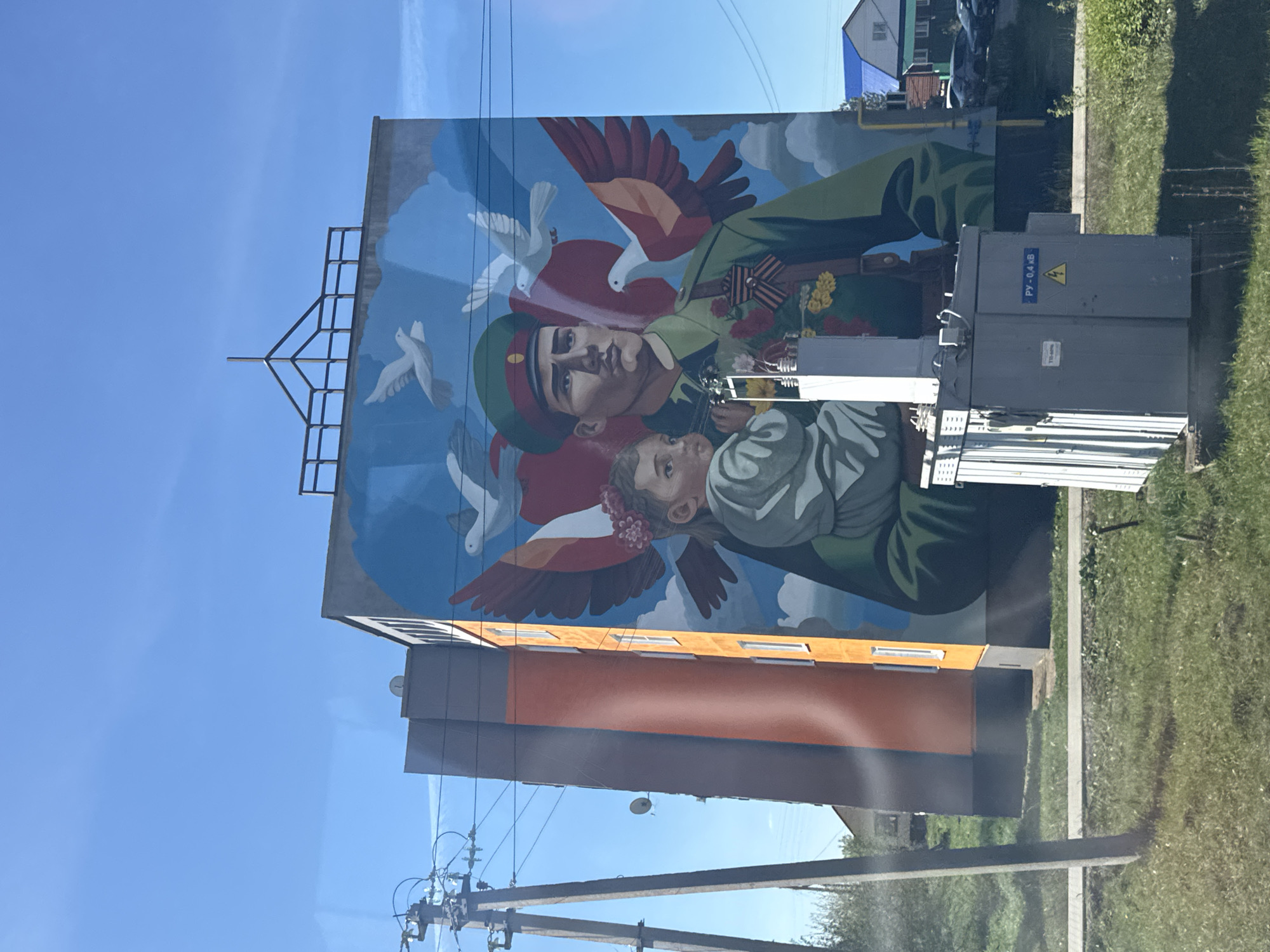 A vibrant mural dominates the side of a building, depicting a soldier in uniform holding a child, surrounded by doves. The soldier wears a green uniform with a red ribbon adorned with flowers, signifying Victory Day celebrations common in Russia. The child, wrapped in a light-colored cloth, gazes upwards. The background consists of a blue sky with white clouds.
The building supporting the mural is painted orange on its upper portion, with a grey section below. An electrical transformer box is attached to the side of the building, partially obscuring the mural. Electrical wires and a wooden pole are in the foreground. A grassy area is visible on the right side of the image, and a blue building can be seen in the background.
The location is likely Osa, Russia, given the theme and artistic style of the mural. The scene appears to be captured during daytime, with clear blue skies.