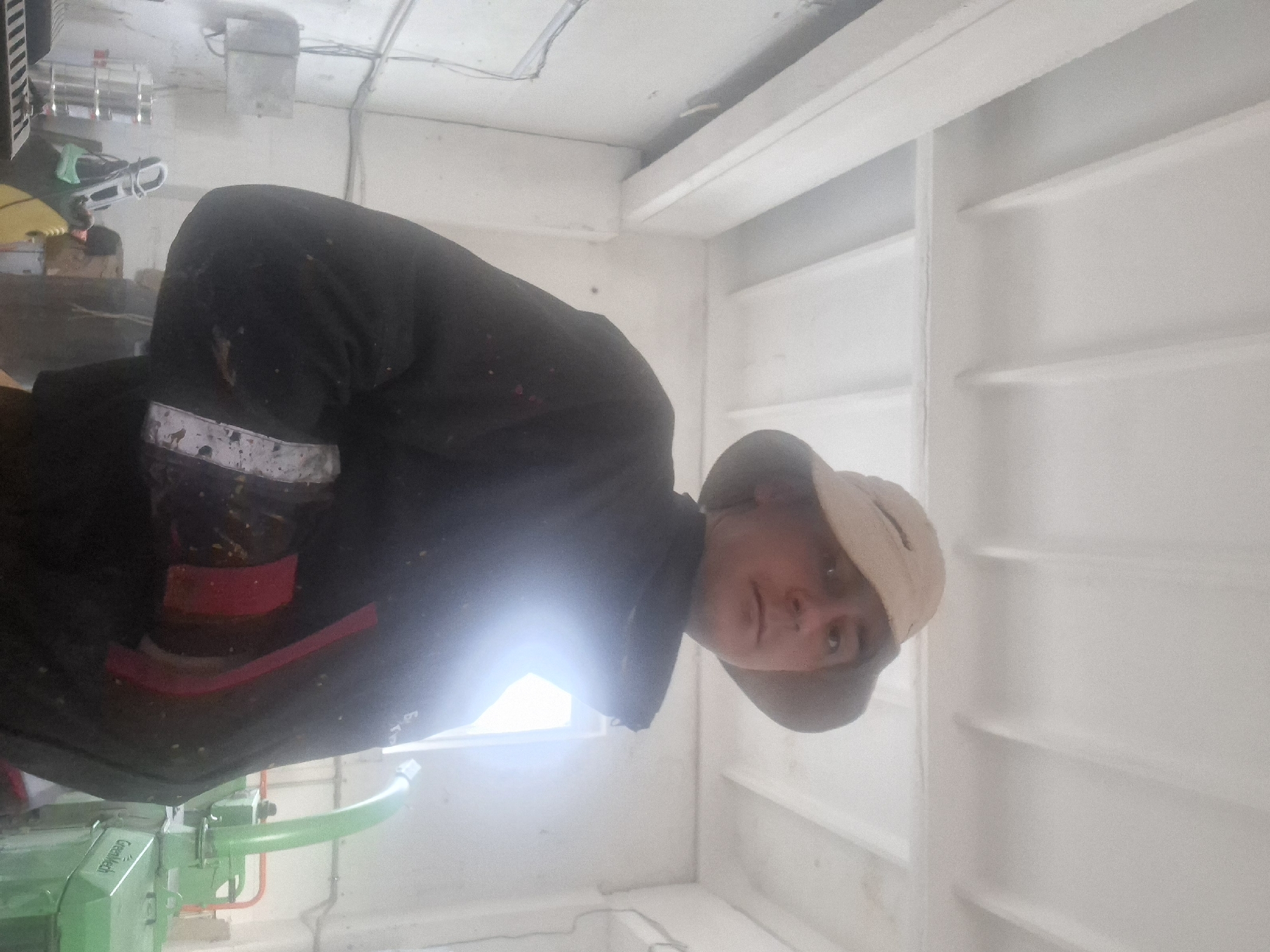 A person wearing a black jacket, a tan hat, and protective gear on their arm stands in a white-walled room, possibly the interior of a boat or building. The room has a grid-like pattern on the walls. A green machine with the text "GreenMech" is visible in the lower-left corner. The location is Unknown, Unknown.