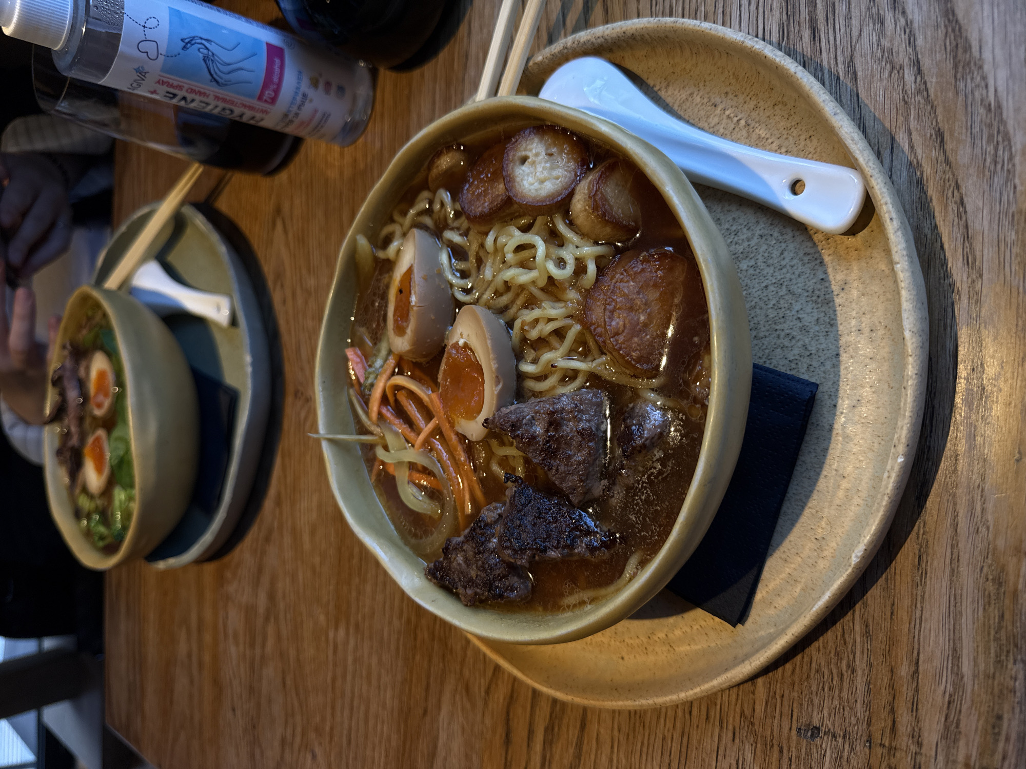 A high-angle shot depicts two bowls of ramen on a wooden table in a restaurant, possibly in Sofia, Bulgaria. The bowl in the foreground, set on a larger plate, contains broth, noodles, sliced sausage, soft-boiled eggs, shredded carrots, and pieces of dark meat. A white ceramic spoon rests on the plate beside the bowl. Chopsticks are positioned across the top of the bowl, and a dark blue napkin is tucked between the bowl and the plate. In the background, another bowl of ramen is visible, featuring similar ingredients. In the upper left corner, there are a bottle of hand sanitizer and other beverage containers. The table has a distinct wood grain pattern. The light suggests an indoor setting.