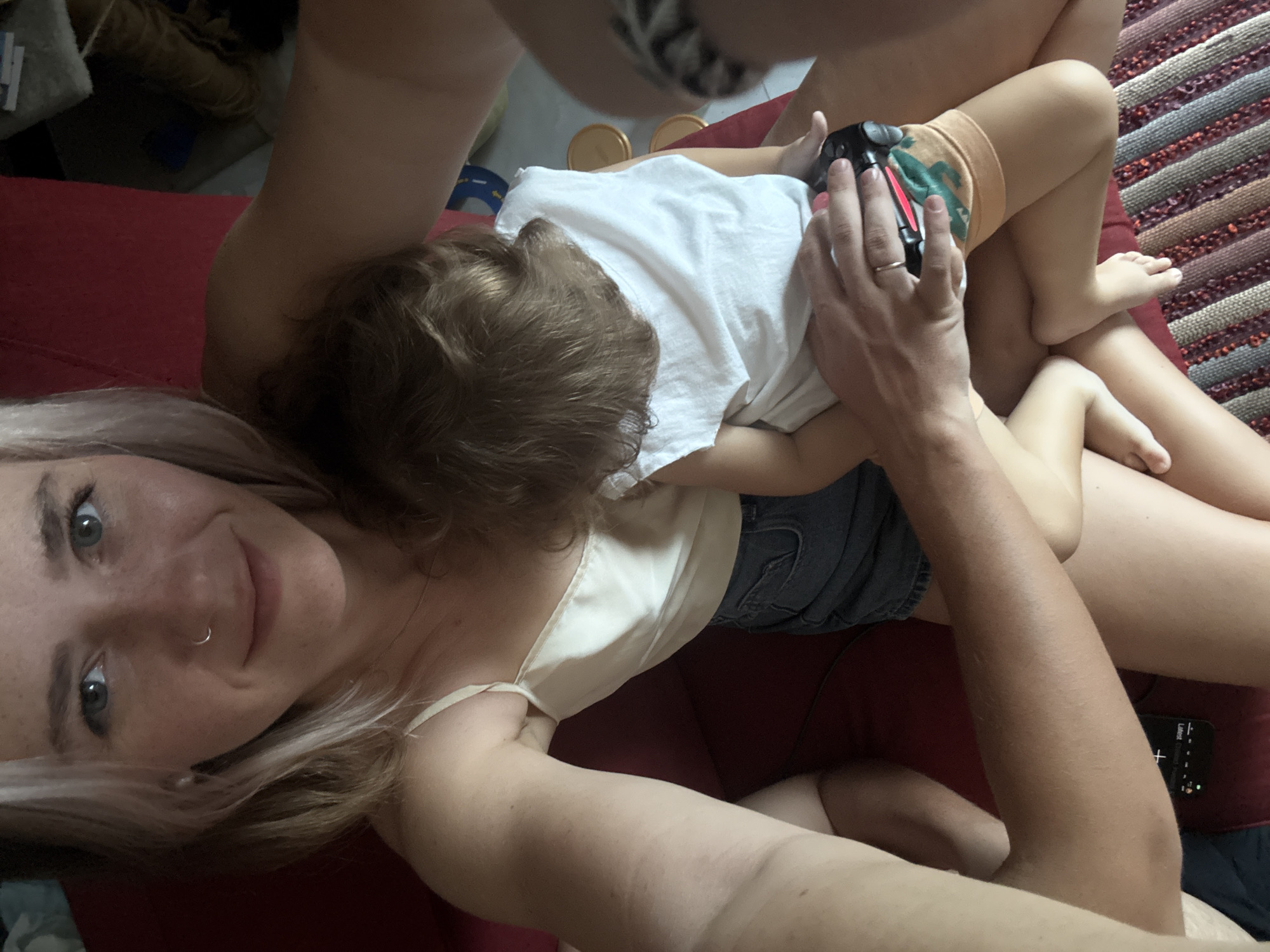A woman with blonde and white hair is holding a young child on her lap. The woman is wearing a white camisole and denim shorts. The child is wearing a white t-shirt and patterned shorts. The woman's left arm is around the child. The child is facing away from the camera, and their head is resting on the woman's chest. The woman's right hand is on the child's leg, and she is holding a video game controller. The background is out of focus, but appears to be indoors with a red couch and a striped rug. The scene suggests a relaxed, domestic setting. The image does not provide specific details to pinpoint the location within Choeng Thale, Thailand, as it lacks any identifying landmarks or contextual elements.