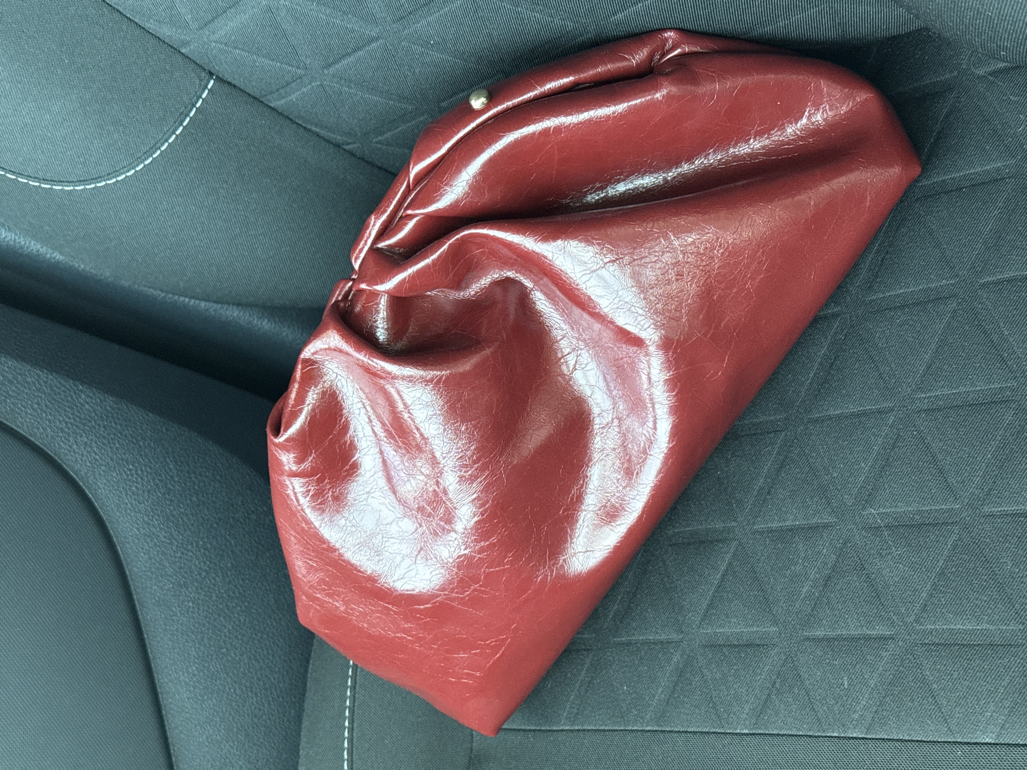 A deep reddish-brown, glossy clutch bag with a textured surface rests on a dark grey fabric car seat. The clutch is gathered at its top opening, displaying a small, round, metallic stud on the upper right side. The car seat features distinct white dashed stitching along one seam and a subtle, embossed geometric diamond pattern across its fabric. The scene is located within a vehicle in the city of Izhevsk, Russia.