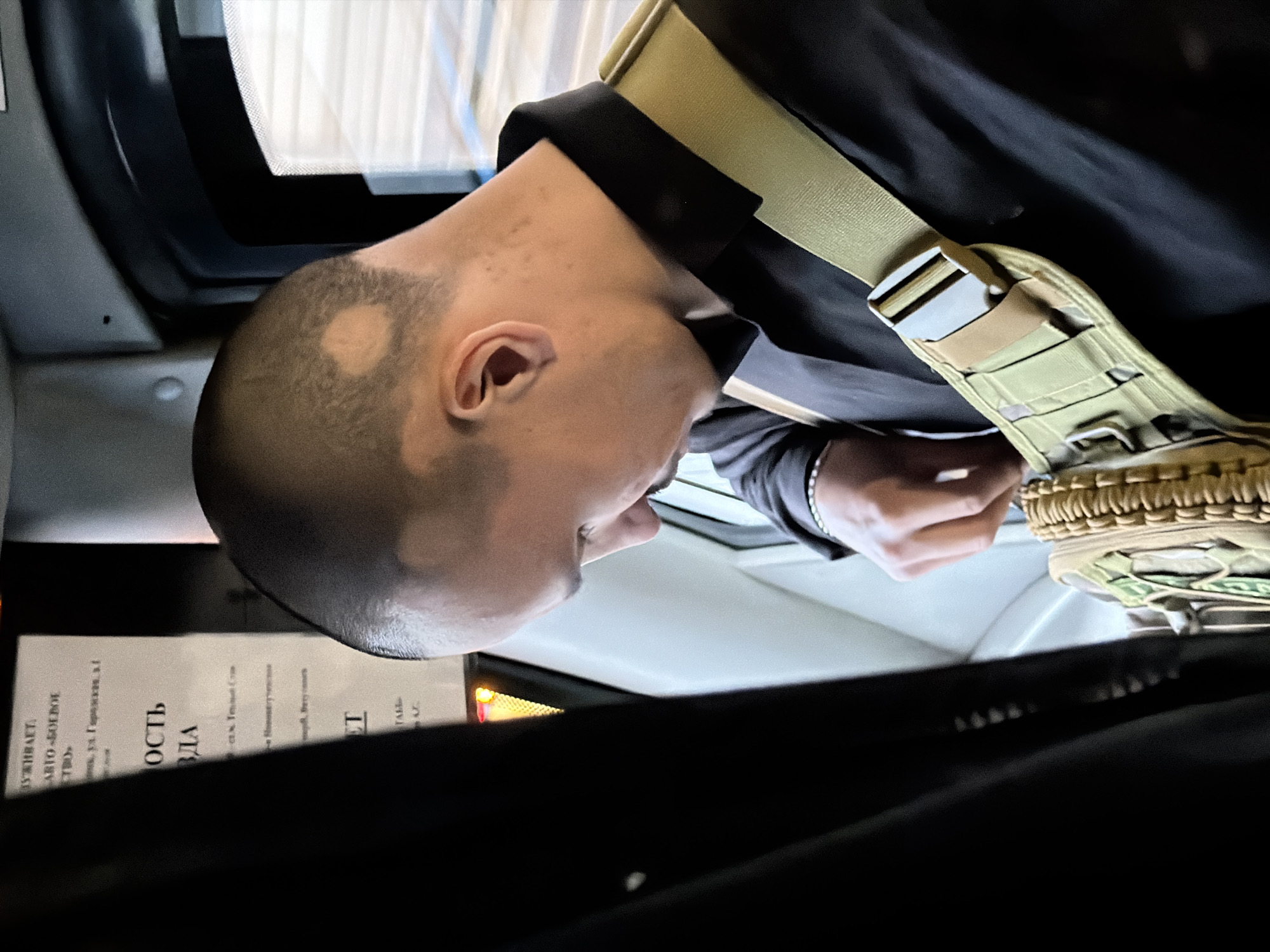 A medium shot from inside a vehicle looking towards the passenger seat, the interior is dark with limited light. A man with a closely shaved head is leaning forward and looking down towards a beige strap with a metal buckle that crosses his shoulder. The man is wearing a black collared shirt. On the head, there is a round patch of hair, likely a result of shaving. In the background, a piece of paper with text written in a language that resembles Russian or Ukrainian is visible. The location is Unknown, Unknown.