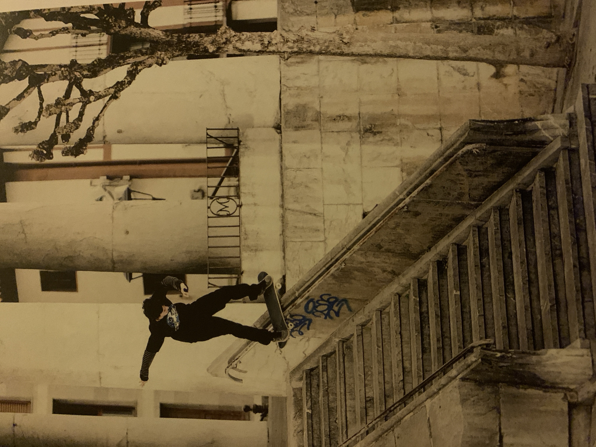 An off-kilter photograph depicts a skateboarder performing a trick near or on a set of concrete stairs adjacent to a building. The location is suggested as Barcelona, Spain, given the style of the architecture, but this is difficult to confirm. A person, dressed in dark clothing and exhibiting a graphic t-shirt or sweatshirt, is positioned mid-air, executing what appears to be a skateboarding maneuver. The person is positioned horizontally, with their body extended and limbs angled. They are wearing striped arm-warmers or gloves.
The skateboard is oriented in a similar plane, slightly above the skateboarder, which suggests they are airborne and engaged in performing a trick.
The background features a set of concrete stairs that leads alongside a multi-story concrete building. The stairs have a concrete railing and are marked with graffiti.
There is a dark-colored tree branch at the top of the frame, which could be a detail of the surrounding park. A fire escape or metal ladder is visible on the side of the building. The building's facade is textured with aging signs, and some windows or recesses.
