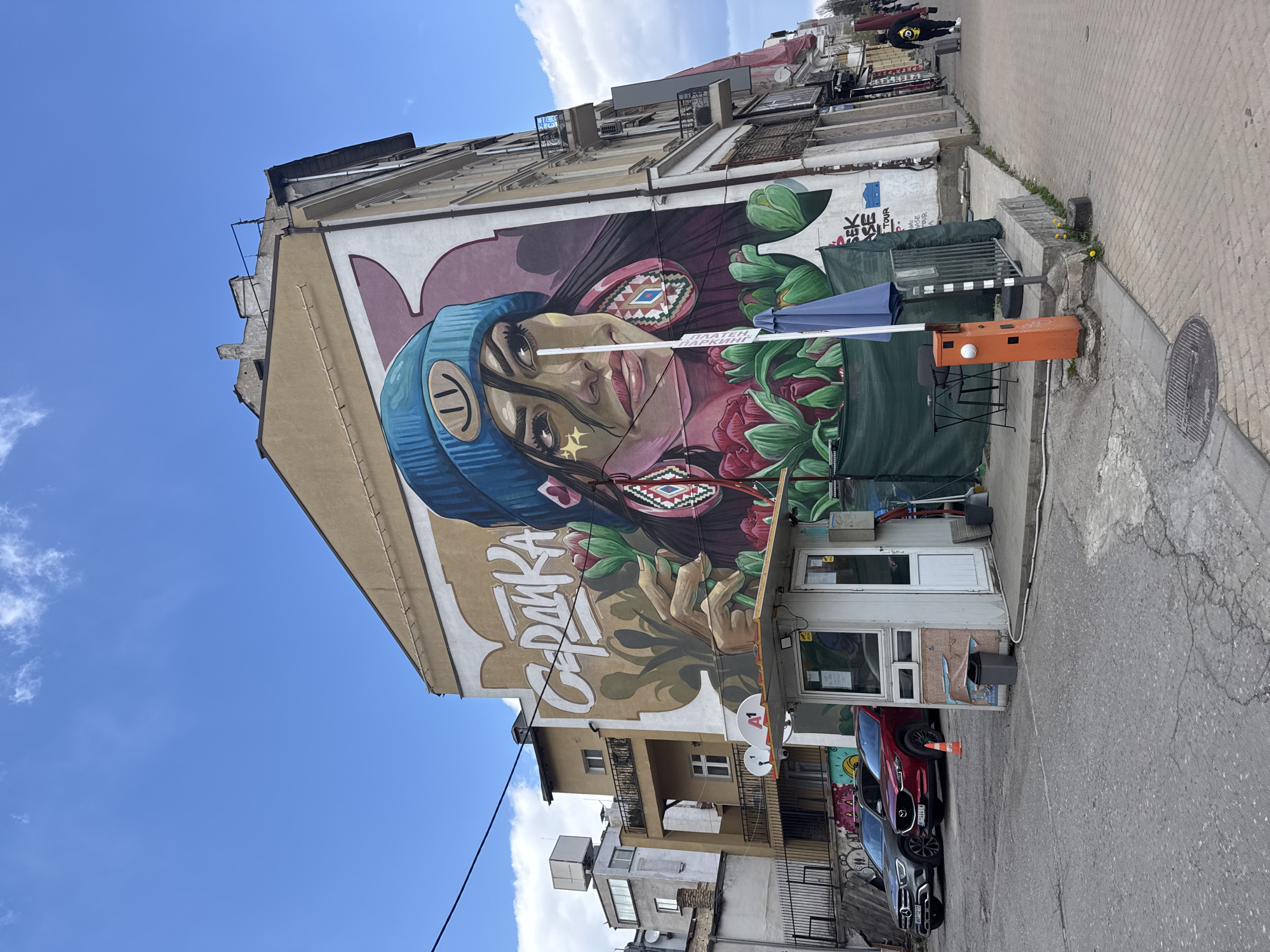 This photograph shows a large mural on the side of a building in Sofia, Bulgaria. The mural depicts a woman with dark hair, wearing a blue beanie with a smiley face. A gold star is painted near her eye, and she is adorned with geometric patterns on her face. She is surrounded by stylized flowers, and the Bulgarian word "САДИКА" is written alongside her image.
The building appears to be several stories high and is situated on a street with other buildings, including cars and street furniture. There is a small structure with a door and windows attached to the building below the mural.