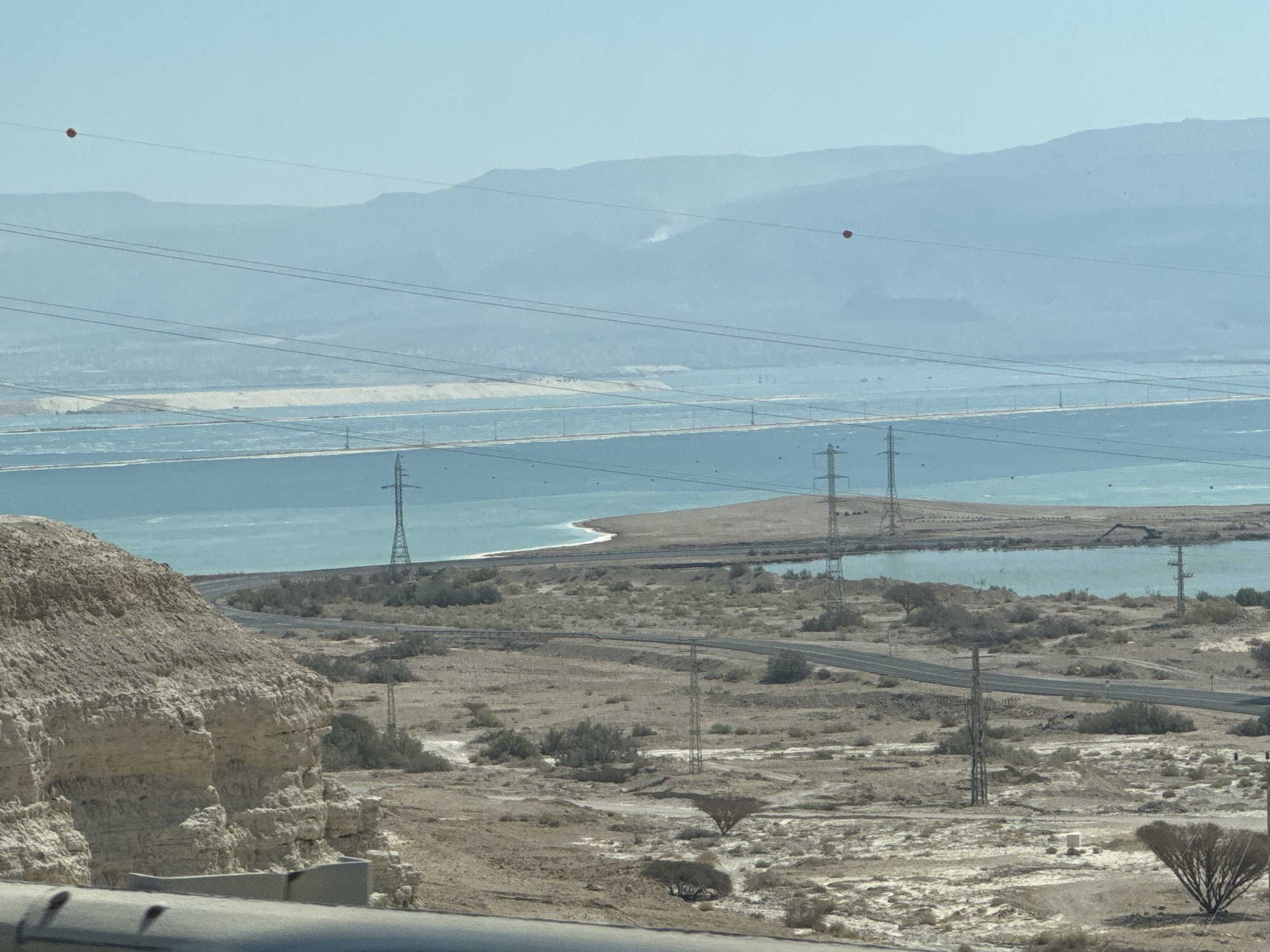 This image depicts a wide, arid landscape with a large body of water in the middle ground, likely the Dead Sea, within the Tamar Regional Council, Israel. In the foreground, a rocky cliff face dominates the left side, with a road curving through the dry, scrub-covered terrain beyond. Multiple tall, metal power line towers are strategically placed throughout the scene, supporting a network of overhead electrical wires that stretch across the frame. Two small, red spherical objects are visible hanging from these wires, positioned against the hazy, blue mountains in the distance. The sky is a clear, pale blue.