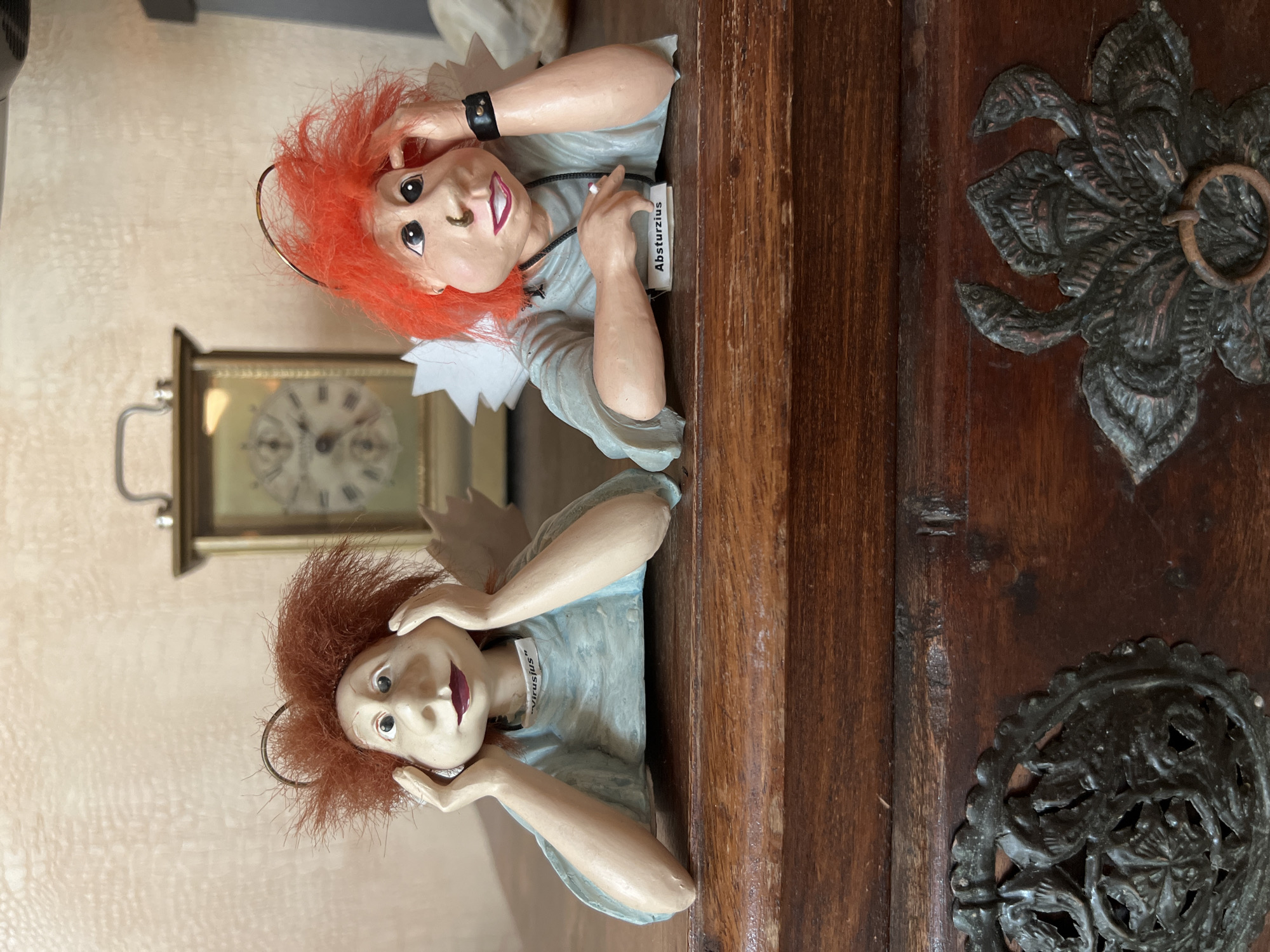 Two whimsical doll-like figures with wild red hair and exaggerated features are perched on a dark wooden surface. The figure on the left has its hands cupping its cheeks and a small white tag on its blue garment reads "Virusjus". The figure on the right has one hand on its forehead, a nose ring, and a black band on its wrist. A small white tag on its blue garment reads "Absturzius". Behind them, on a lighter textured wall, is a vintage-style alarm clock with Roman numerals. The wooden surface appears to be the lid of a chest, adorned with ornate dark metal embellishments. The scene is brightly lit, highlighting the details of the dolls and the chest. The location is not specified in Bender, Moldova.