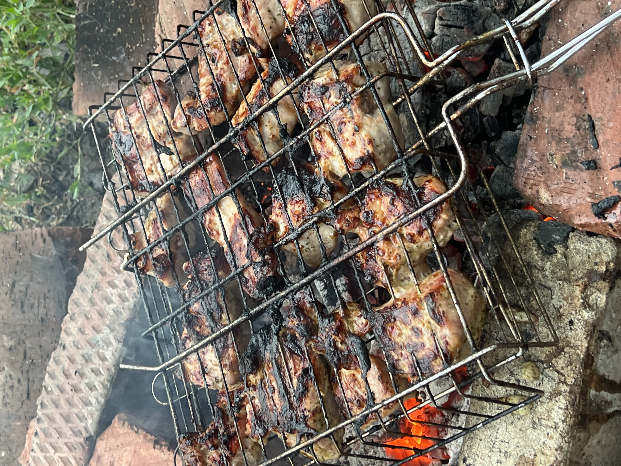 This image depicts a close-up, overhead view of meat cooking on a metal grill basket over hot coals. The meat appears to be chicken or pork, with some pieces showing charring and browning from the grill. Smoke is visible rising from the meat and coals. The grill is situated within a makeshift fire pit constructed from bricks and rocks. The background shows more bricks and some blurred green foliage, suggesting an outdoor setting. The scene is consistent with a barbecue or outdoor cooking event. There are no people visible in the image. The location in Kamen-na-Obi, Russia, is inferred from the provided context and is not visually discernible within the image itself.