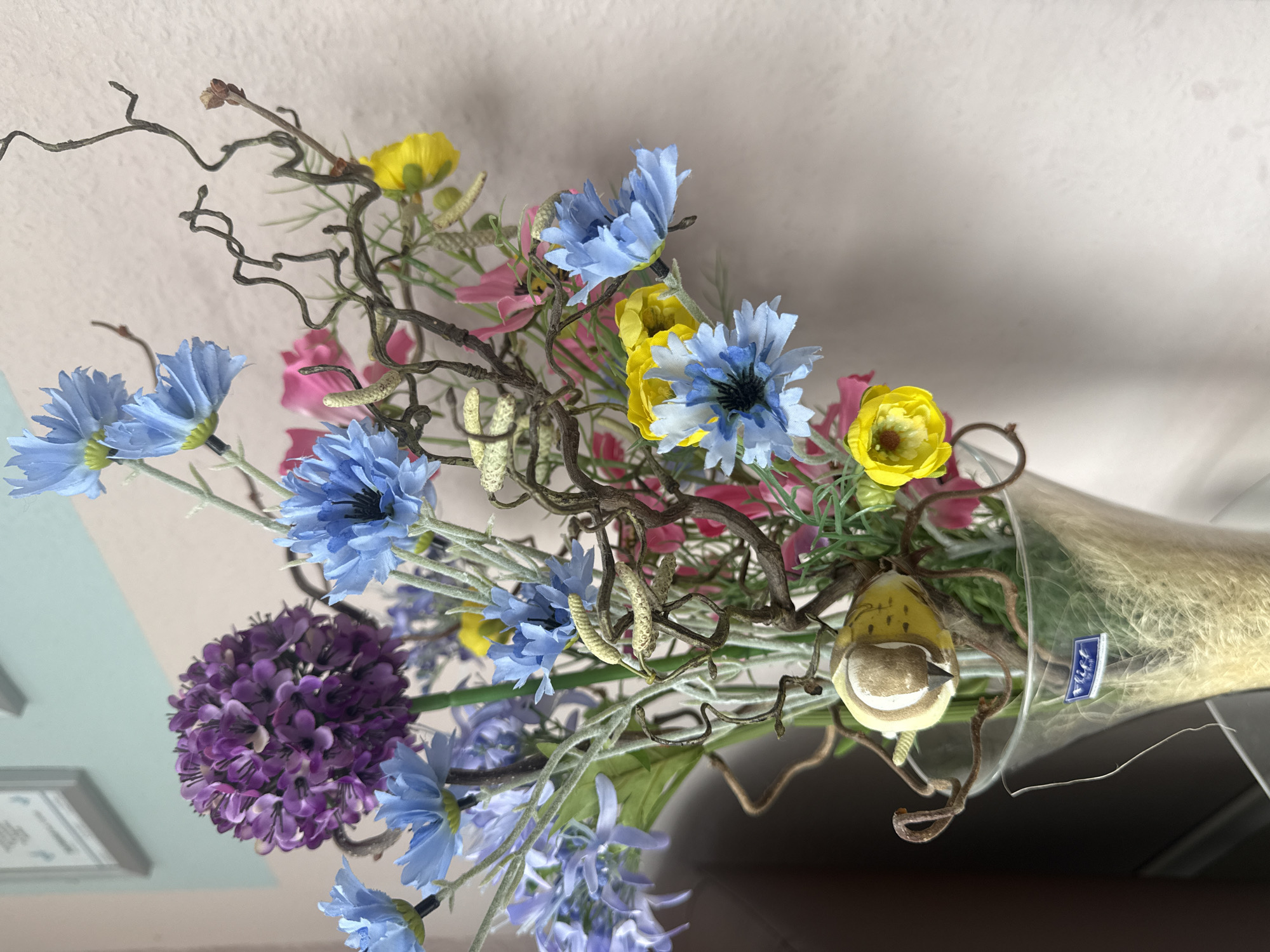 A still life arrangement features a clear glass vase filled with artificial flowers and decorative branches. The vase contains a mix of blue, yellow, and purple flowers, along with twisted brown branches and some light-colored filler material. A decorative bird-like ornament is positioned among the branches. The vase itself has a blue sticker with white text, possibly a brand label. The background consists of a textured, light-colored wall. The scene is indoors, with a hint of a darker object, possibly furniture, on the left side and a framed picture on the wall in the upper left corner. The lighting suggests it is daytime. This image depicts an indoor decorative arrangement, not a scene in Heiligenrode, Germany.