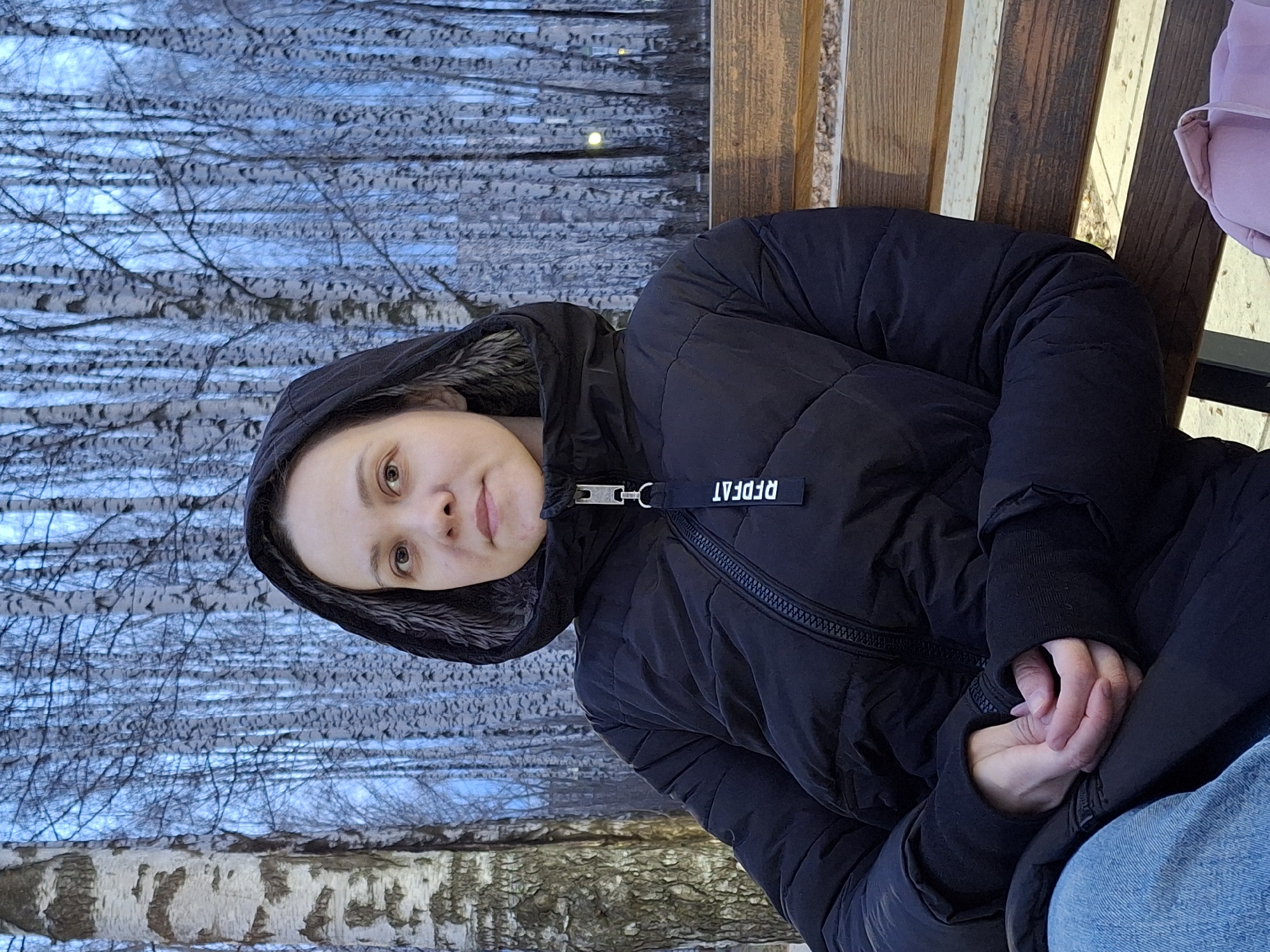 The image captures a young woman seated on a wooden bench outdoors in Chaikovsky, Russia. She is wearing a black padded winter jacket with a fur-lined hood and blue jeans. Her hands are clasped together in her lap, and she looks directly at the viewer with a neutral, calm expression. Her eyes are light brown.
The setting is a park or forested area, characterized by a dense background of bare birch trees, their white and black bark prominent against a cool, possibly overcast, sky. The absence of leaves on the trees suggests a cold season, likely winter or early spring, consistent with the heavy jacket worn by the subject.
A distinct black fabric tag is attached to the zipper of her jacket, displaying white text that reads "RFDFAT," which appears to be a stylized or reversed version of "REPEAT." Part of a light purple bag or item is visible on the bench beside her. The overall atmosphere is quiet and serene, with soft daylight illuminating the scene.
