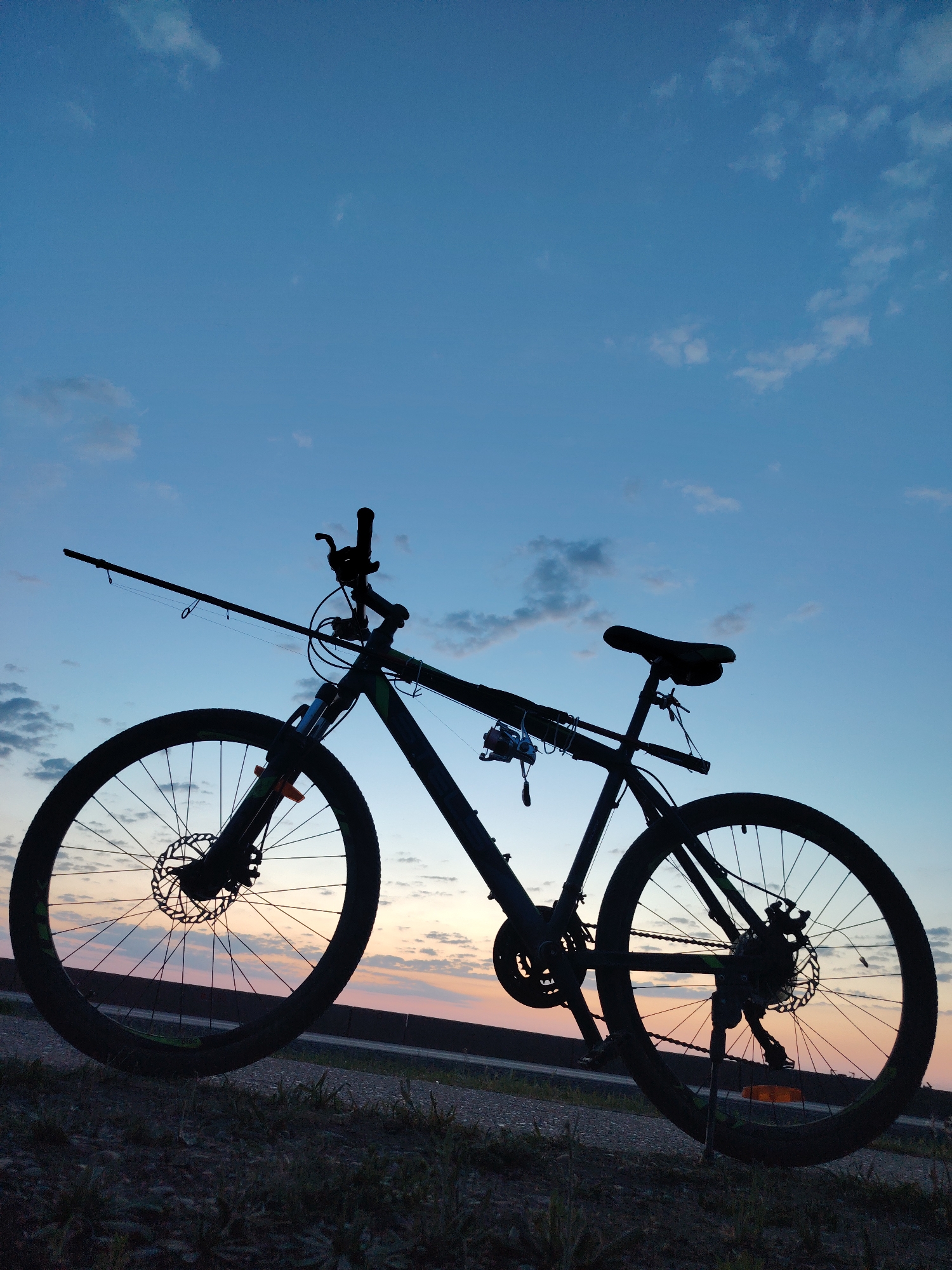 The image depicts a bicycle silhouette against a dusk sky. A fishing rod is attached to the bicycle frame. The background includes a road or path and a sky with a gradient of colors. The location is Chaikovsky, Russia.