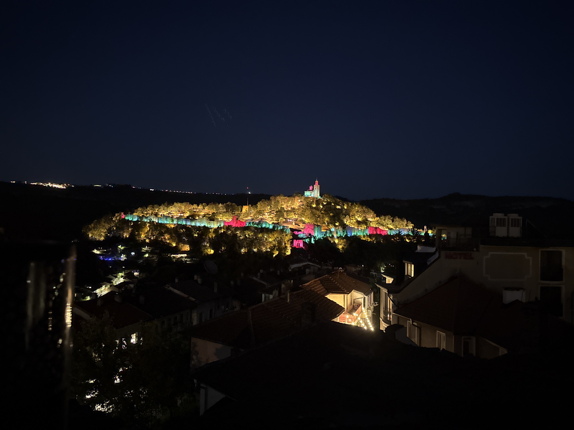 This is a night-time, high-angle, medium-shot photograph of the Tsarevets Fortress in Veliko Tarnovo, Bulgaria. The fortress is illuminated with multi-colored lights, predominantly red and green, with a prominent red-roofed tower visible on the peak. Below the fortress, a city sprawls out, with buildings illuminated by warm indoor lighting and some decorative string lights visible on a balcony. The sky is dark, with faint white trails possibly from an airplane or distant contrails. The foreground is dark, with shadows obscuring most details, suggesting the photo was taken from a balcony or elevated vantage point. There are no people or animals visible in the scene. The overall impression is of a vibrant, illuminated historical landmark overlooking a dark, quiet city at night.