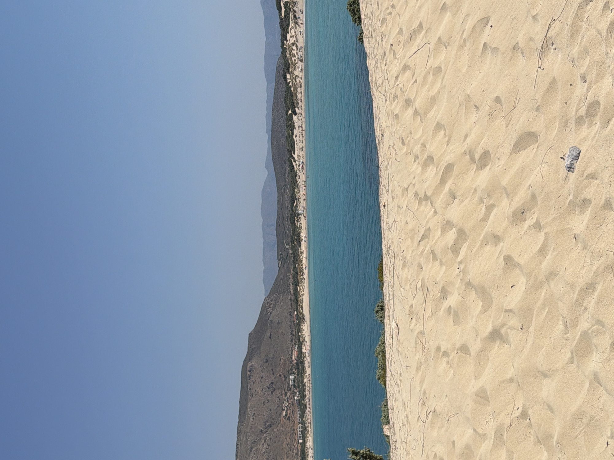 The image shows a sandy beach in Greece. The foreground is dominated by sand dunes, with some dry vegetation scattered across the surface. In the midground, a body of turquoise water stretches out, with a shoreline visible in the distance. The shoreline is lined with sand dunes and dotted with small buildings, suggesting a coastal settlement. Beyond the settlement, a range of low, barren hills forms the background, meeting a clear, pale blue sky. The scene is bathed in bright sunlight, indicating a clear day. There are a few people visible on the beach in the distance, and some beach umbrellas.