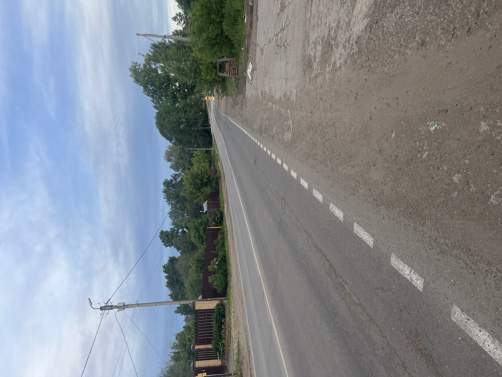 The image depicts a road in the city of Chaikovsky, Russia. It is a paved road with white dashed lane markings. The road appears to be mostly straight. The right side of the road has a wide shoulder with gravel and some debris. On the left side, a fence and trees border the road. Utility poles with wires are visible along the left side of the road. The sky is partly cloudy with a light blue color. The image appears to have been taken during daylight hours.
