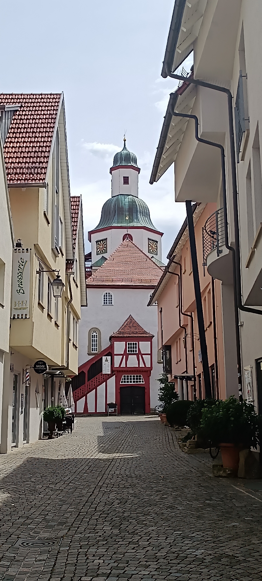 The image depicts a cobblestone street in Winnenden, Germany, leading towards a church with a prominent tower. The street is lined with buildings on both sides. On the left, there is a yellow building with red-tiled roofs, featuring a sign that reads "Brasserie" and "B-E-N-Z," as well as a barber shop sign for "MORI Friseursalon." Further down the left side, outdoor seating is visible.
The central focus is the church, characterized by its white facade, a red-tiled roof, and a distinctive tower with a green dome. The tower has two visible clock faces. Adjacent to the church, on the right side of the street, is a smaller, red and white half-timbered structure with an external staircase leading up to a red-tiled roof.
On the right side of the street, the buildings are painted in lighter colors, including pink and white. There are balconies and gutters visible on these structures. Several potted plants are placed along the street on both sides, and shadows from the buildings cast across the cobblestones. The sky is overcast with clouds. No people are visible in the scene.