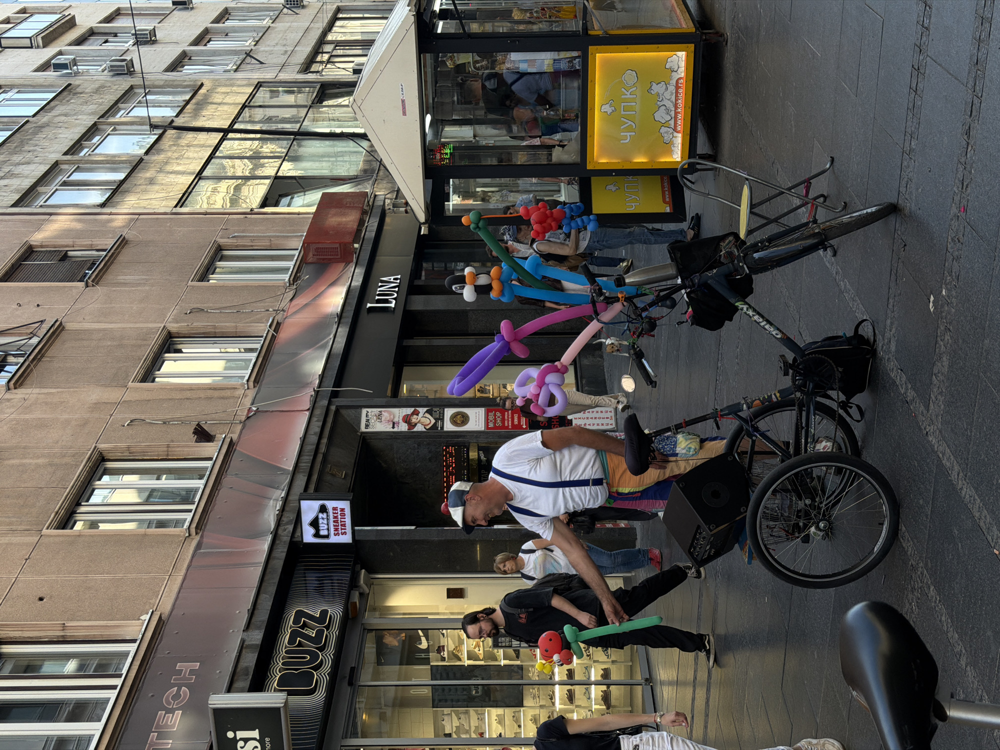 This image shows a street scene in Belgrade, Serbia. Several people are present, including a balloon artist wearing a white shirt, suspenders, and a cap, as well as other people walking by or standing near storefronts. One man is holding a green balloon. A bicycle with various colorful balloons attached to it is parked nearby. The storefronts visible include "BUZZ SNEAKER STATION", "LUNA", and a yellow booth that says "ЧУПКО" (Chupko), a local snack stand. The buildings in the background have multiple windows and air conditioning units. The ground is paved with gray stones.