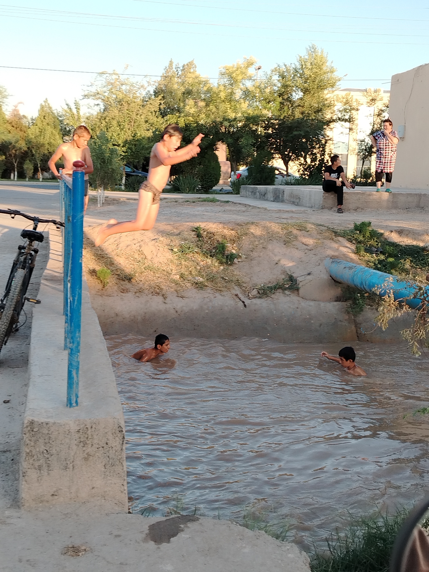 In Khiva, Uzbekistan, on a sunny day, four young boys are engaged in playful activities near a body of water. In the foreground, a bicycle leans against a blue railing. The water appears to be a canal or ditch, with one boy in the water and two others poised to jump or play. One boy is mid-air, jumping into the water, while another stands on the bank, observing. In the background, two women are standing and talking. The environment is characterized by trees, buildings, and a clear sky, suggesting an outdoor urban setting.