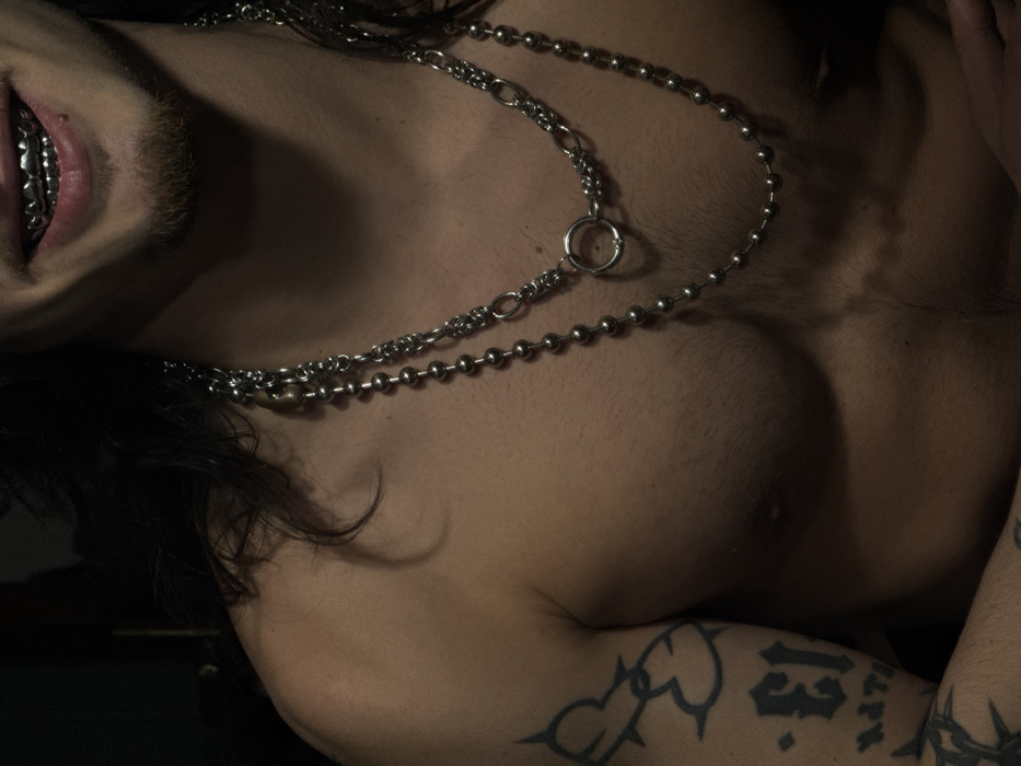 The image presents a close-up, mid-torso to chin-level view of an individual. The subject has long, dark hair and a short goatee beard. A smile reveals silver-colored grillz covering the upper teeth. The individual is shirtless, exposing the bare chest and upper abdomen. Two silver-colored necklaces are worn: an inner chain-link necklace featuring a prominent O-ring pendant, and a longer, outer necklace composed of uniform spherical beads. The left arm is visible, displaying several tattoos, including a barbed wire heart design on the bicep, a stylized numeral "13," and other indistinct script or symbols. The background is uniformly dark and featureless, suggesting an indoor environment with focused lighting on the subject. No specific environmental details within the image allow for the determination of a precise location in the city of Sofia, Bulgaria.