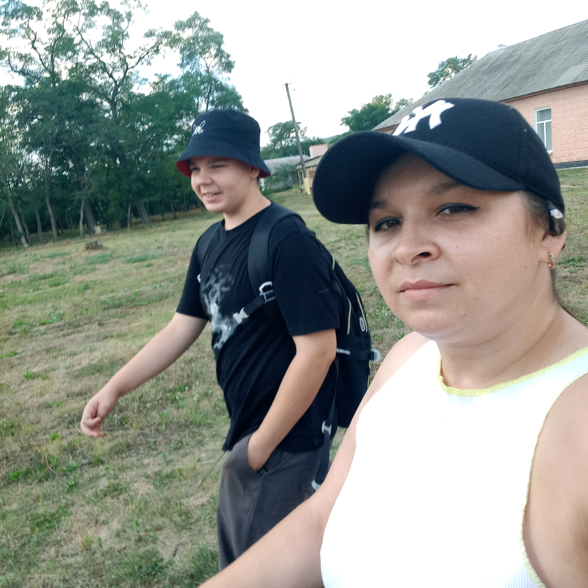 Two individuals, an adult female and a juvenile male, are pictured outdoors on a dry, grassy area. The juvenile male, on the left, is wearing a black bucket hat with a white emblem, a black t-shirt with a graphic design, and dark pants. He has his left hand in his pocket and appears to be walking. The adult female, on the right, is in the foreground and is mostly out of focus. She is wearing a black baseball cap with a white "NY" logo and a white tank top with yellow trim. The background shows a building with a light pink facade and a grey roof, surrounded by trees and a utility pole. The scene suggests a casual outdoor setting. The location is identified as Klyntsi, Ukraine.
