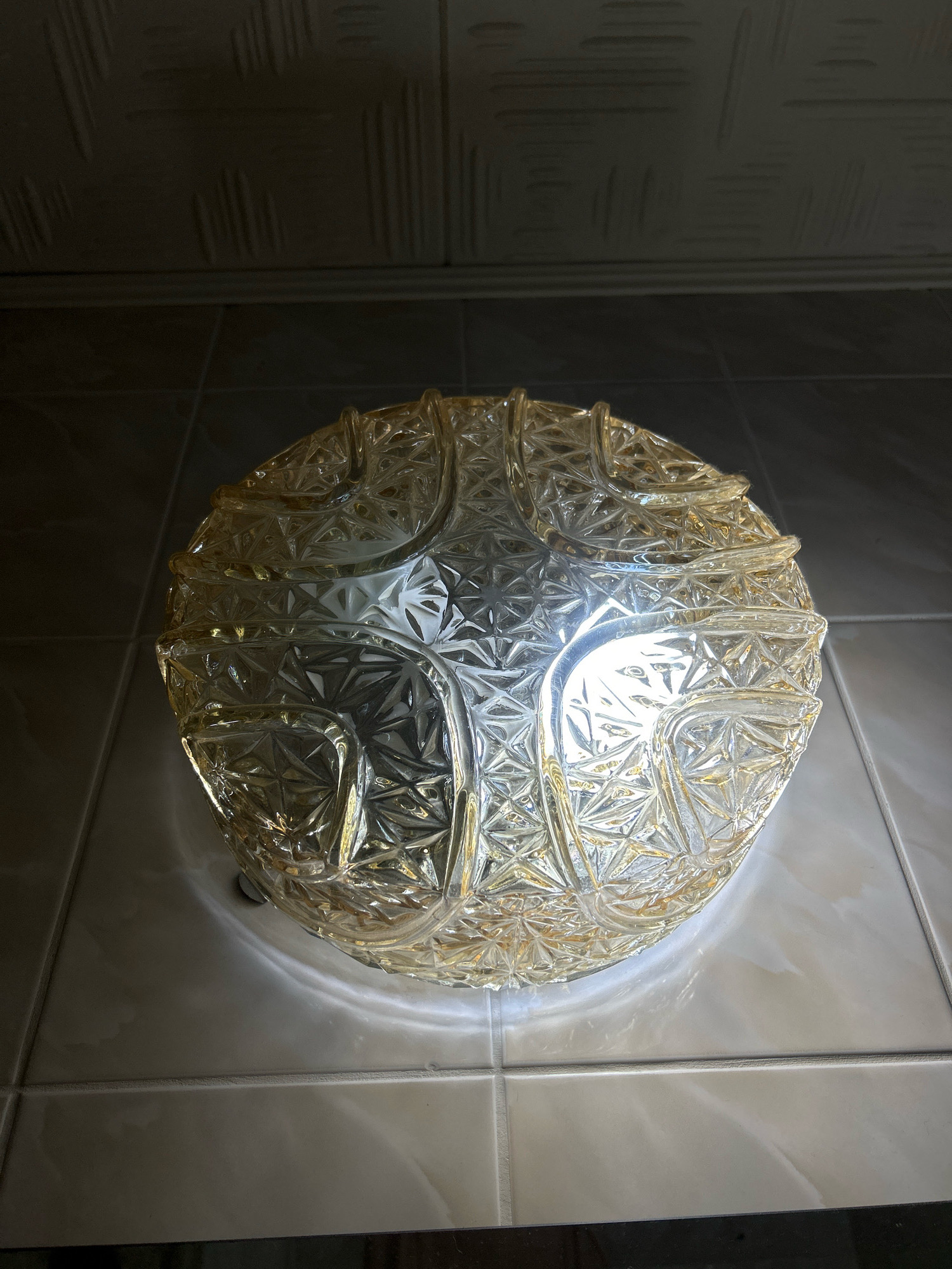 The image shows a close-up, overhead shot of a decorative, spherical glass light fixture with a textured, geometric pattern. The fixture is translucent and appears to be made of a yellowish-tinted glass. It is mounted on a tiled surface, possibly a wall or ceiling, with light-colored square tiles and darker grout lines. Above the fixture, a darker, textured surface is visible, possibly a textured ceiling or a different wall section. The lighting is dim, with the light fixture itself being the primary source of illumination, casting reflections and highlights on the surrounding tiles. The image appears to have been taken indoors. There are no people, animals, or any other objects clearly visible in the scene. There is no information available to suggest this image is located in Moscow, Russia.